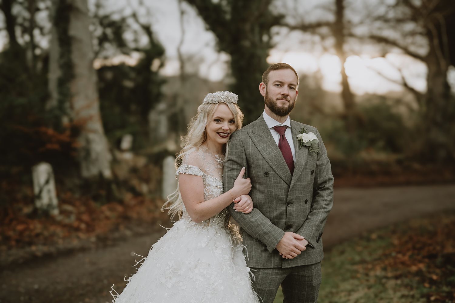 A bride in a white lace gown and groom in a grey tweed suit pose together outdoors at a woodland wedding.