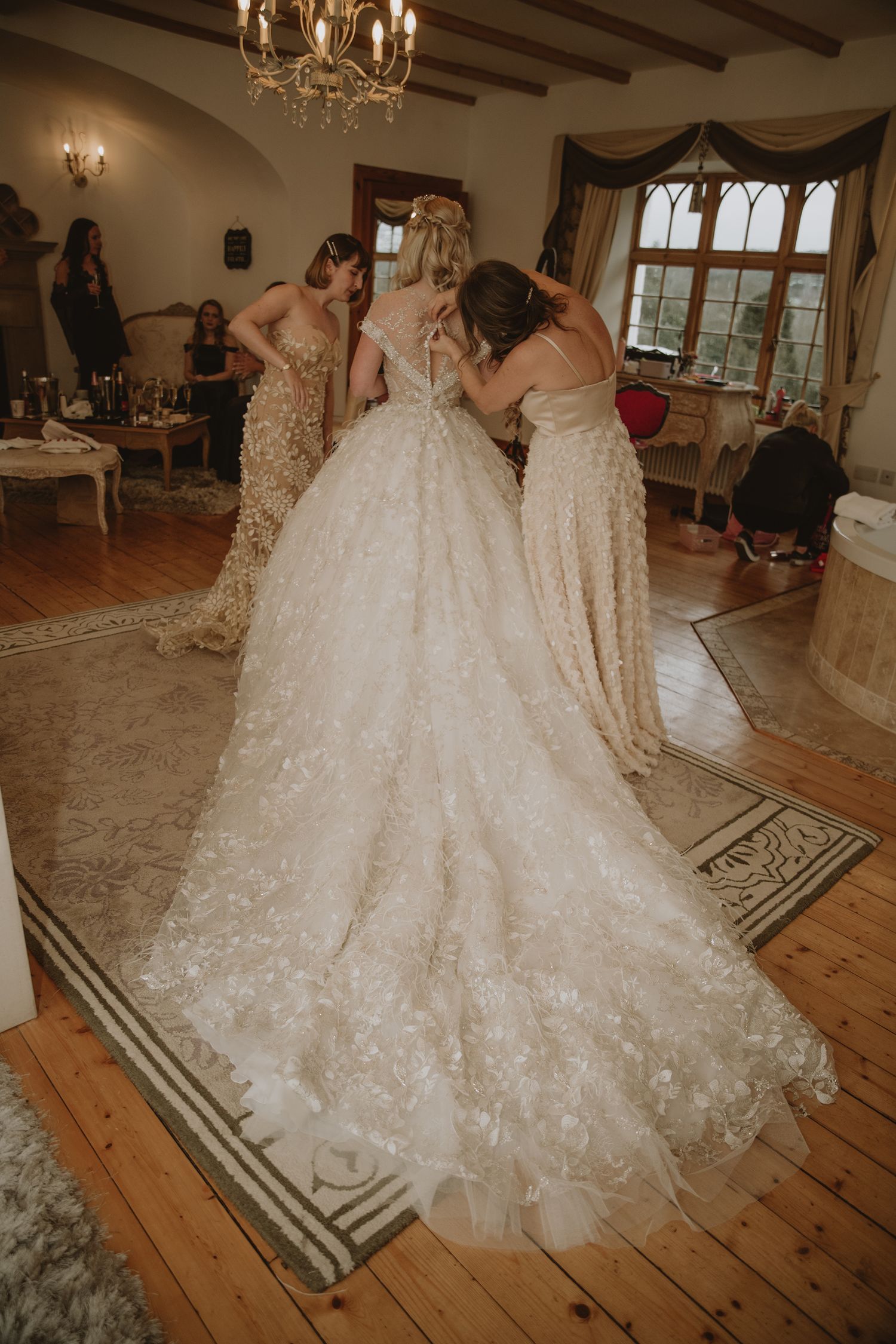 Bridesmaids helping bride in floral ballgown wedding dress with long train in elegant room.