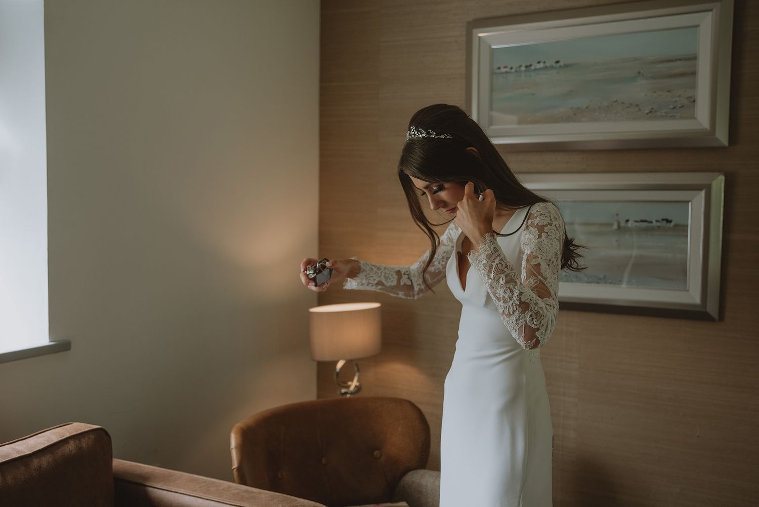 Bride in elegant lace wedding dress adjusting earrings beside a lamp in a warmly lit hotel room.