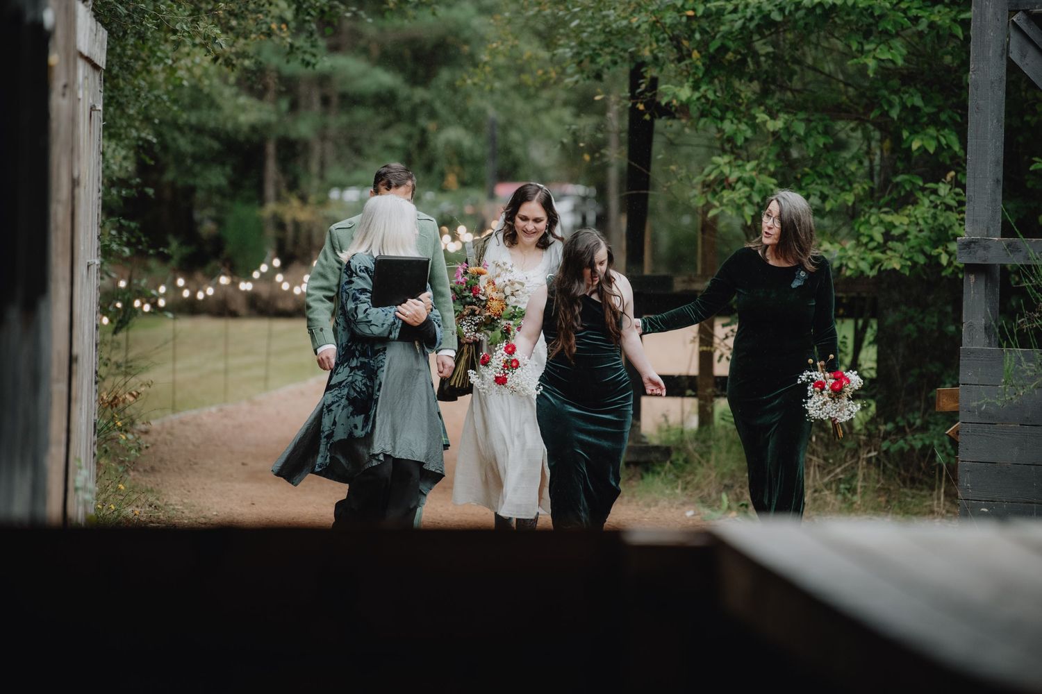 Smiling bride in white gown with bouquet walking with wedding party along a forest path with fairy lights.