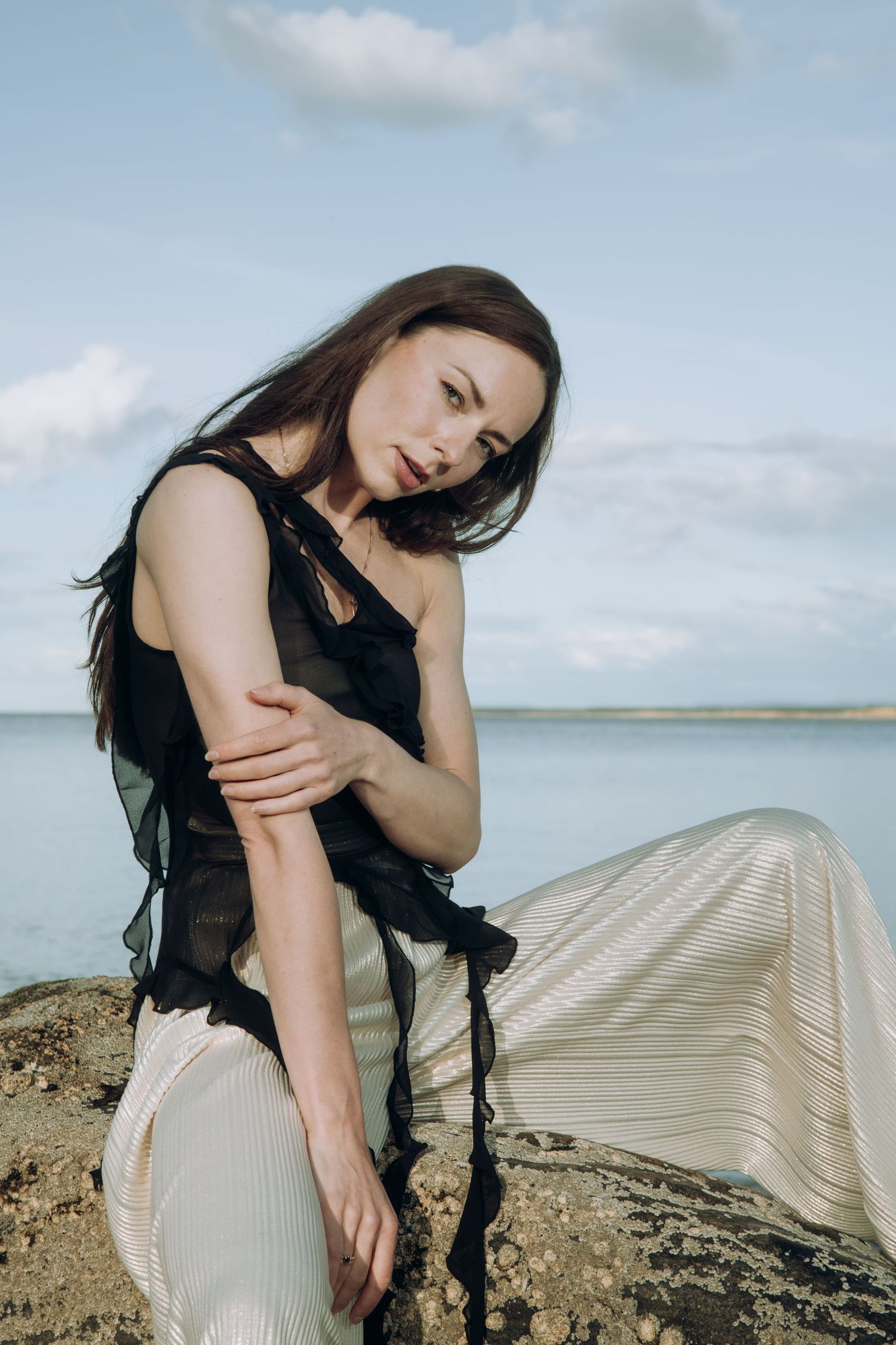 Woman in black sheer top and silver skirt sitting on rocks with blue ocean behind her.