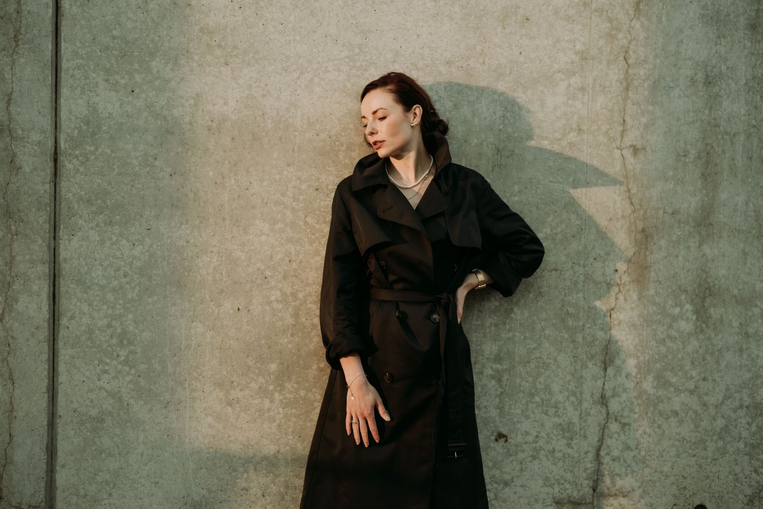 Woman in dark brown belted trench coat poses with eyes closed against a concrete wall.