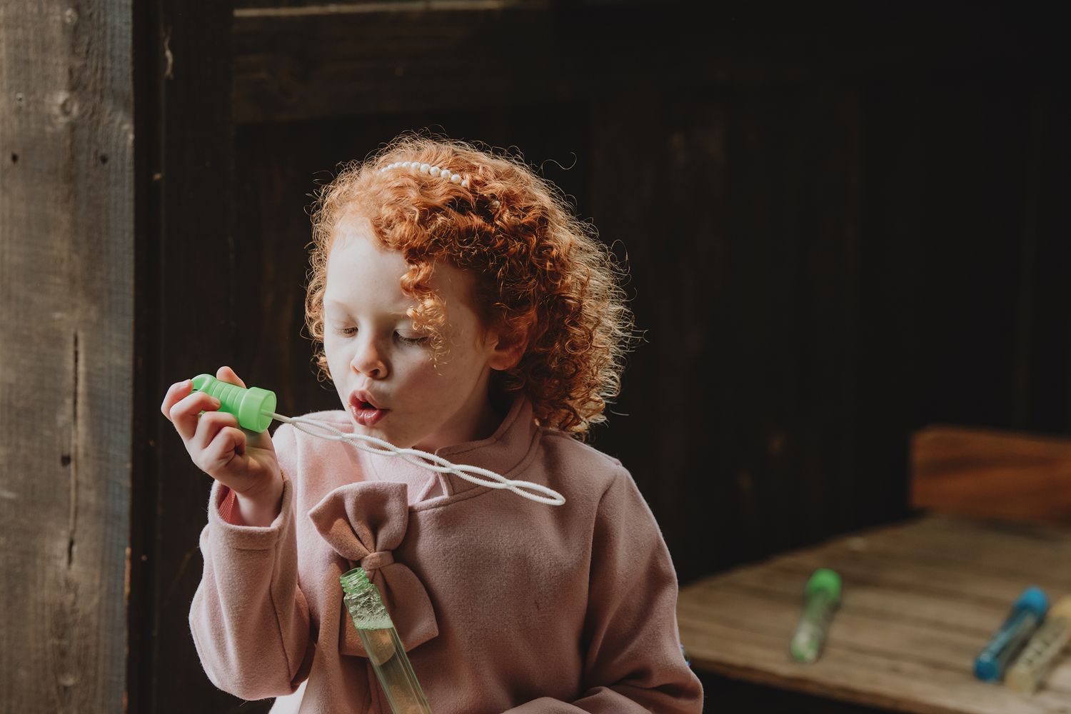 A curly red-haired toddler blowing bubbles outdoors, wearing a pink hoodie.