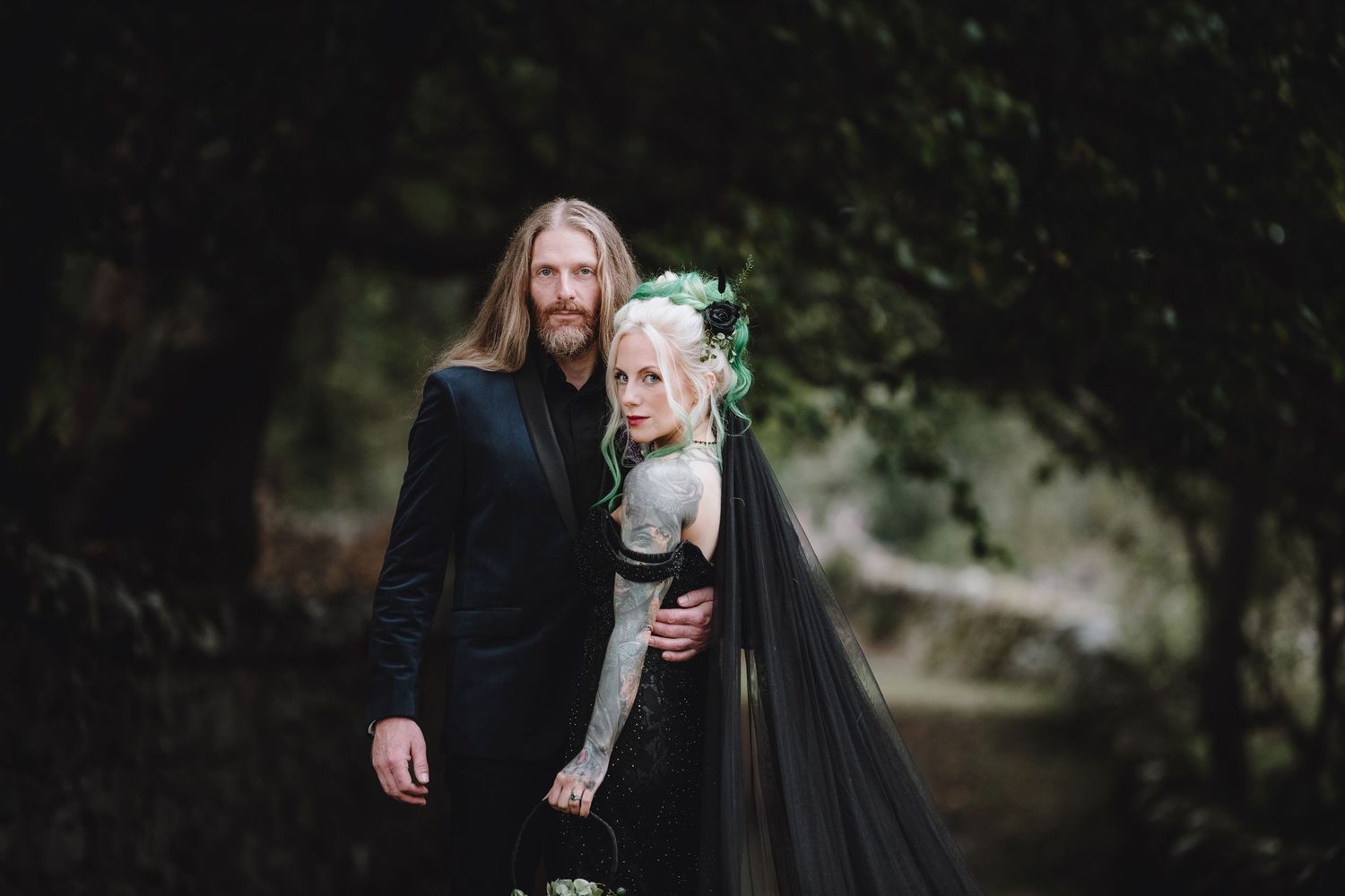 Couple posing in a dark forest, groom in a black suit and tattooed bride in a black wedding dress with green floral crown.