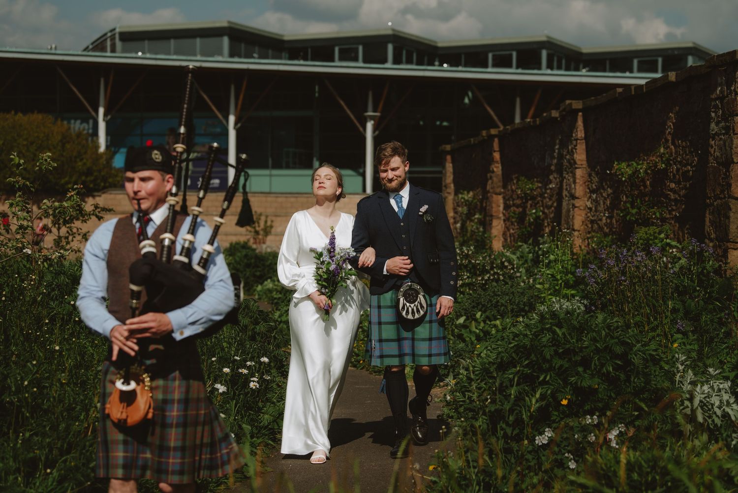 Colorful photo of newlyweds following bagpiper through garden at Scottish wedding venue.