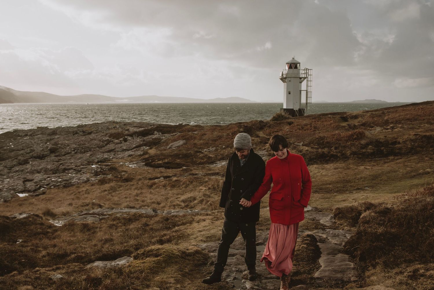 Couple holding hands walking along rocky coastal path with white lighthouse and stormy sea in background.