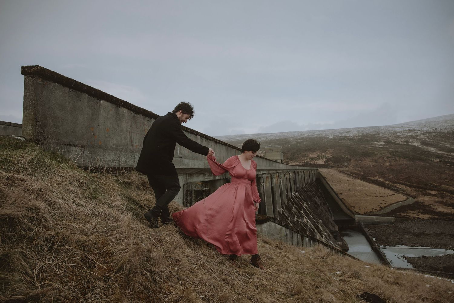 Couple in dramatic attire navigate a misty hillside, woman in flowing red gown guided by partner in black near rustic bridge.