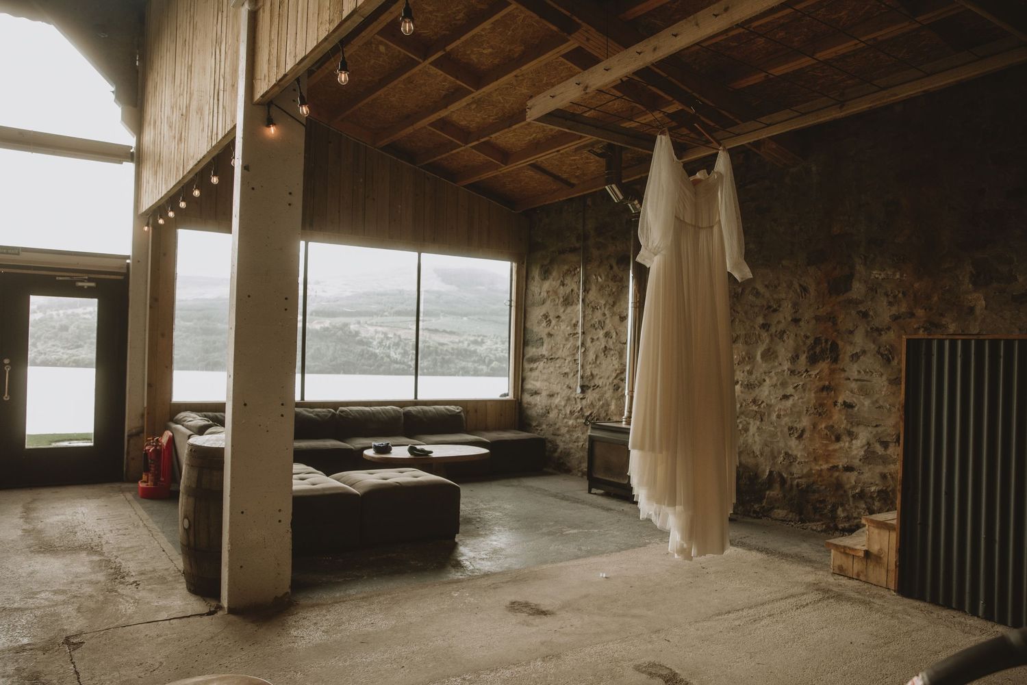 Wedding dress hanging in a rustic stone venue with large windows overlooking a scenic loch and hillside landscape.