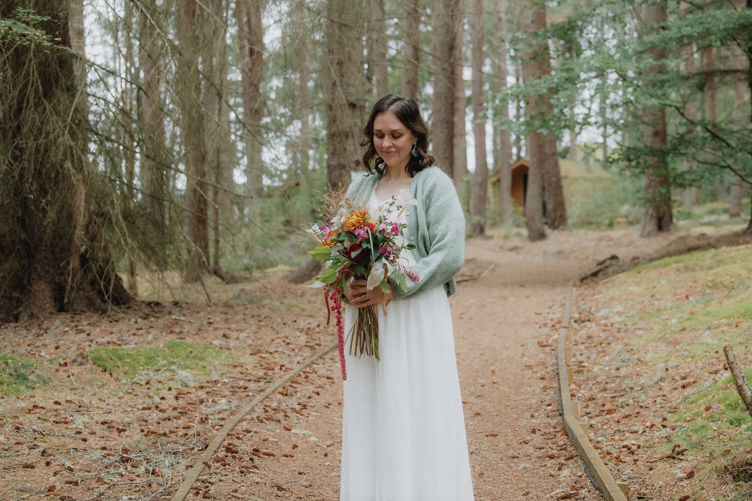 Bride in white dress and mint cardigan smiling at vibrant autumn bouquet in a serene pine forest.