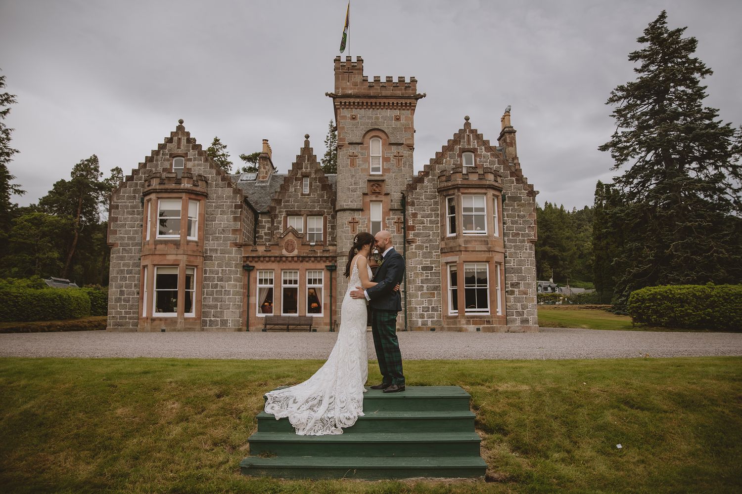 Bride and groom pose romantically on green steps before a stunning Scottish baronial castle on a cloudy day.