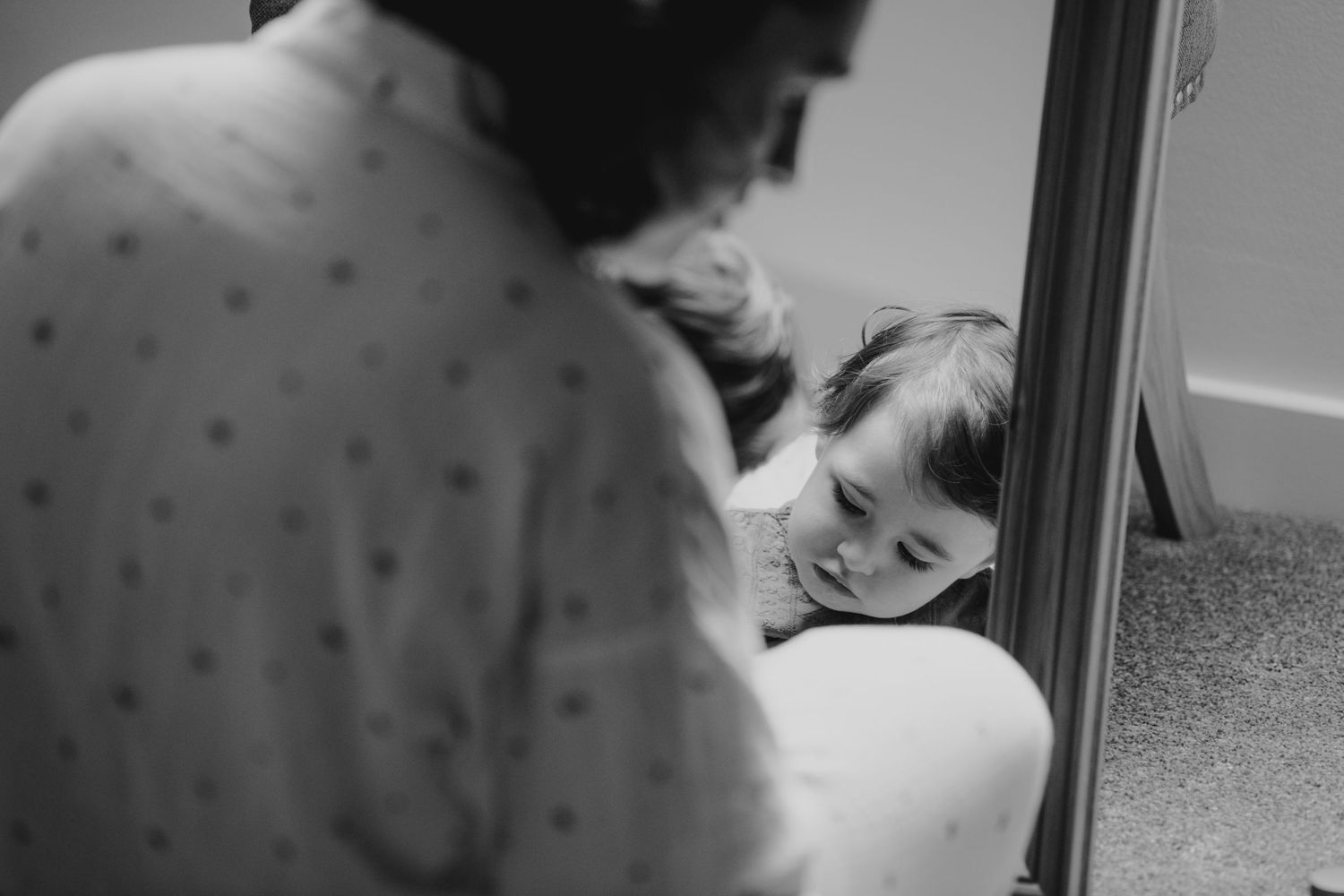 Black and white close-up of a smiling child reflected in a mirror beside a bride getting ready.