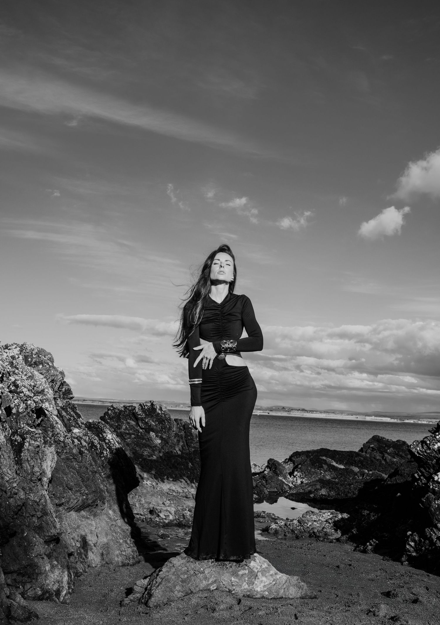 Black and white photo of a woman in black gown standing on coastal rocks under cloudy sky.