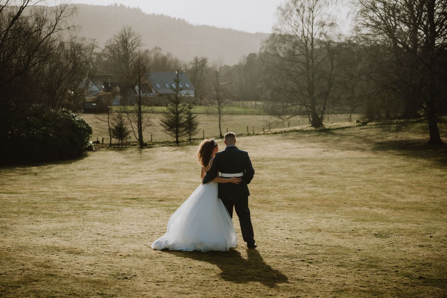 Bride in flowing white gown and groom walk hand in hand across golden sunlit countryside landscape.