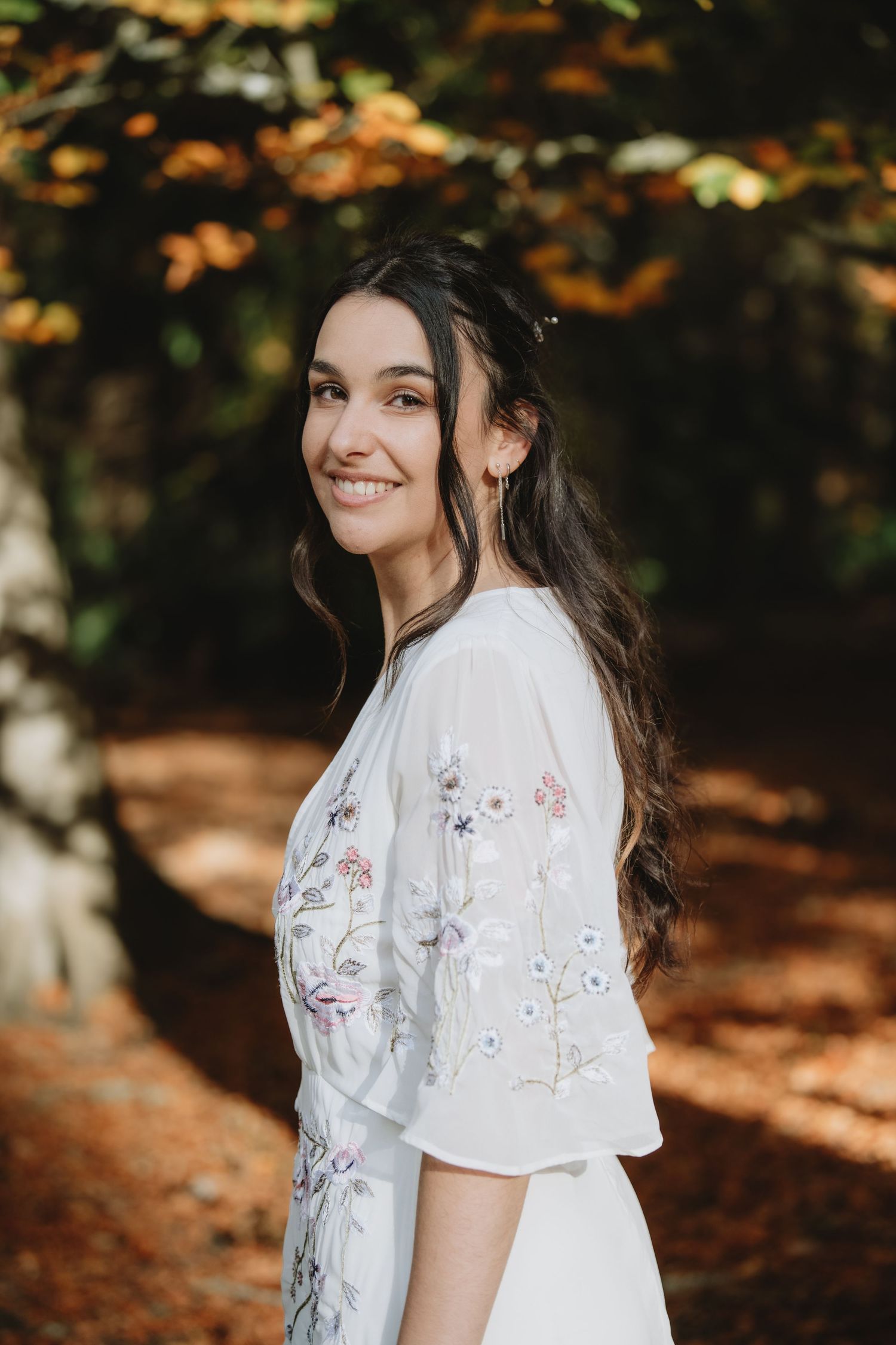 Smiling bride in floral embroidered white dress poses in sun-dappled autumn woodland setting.