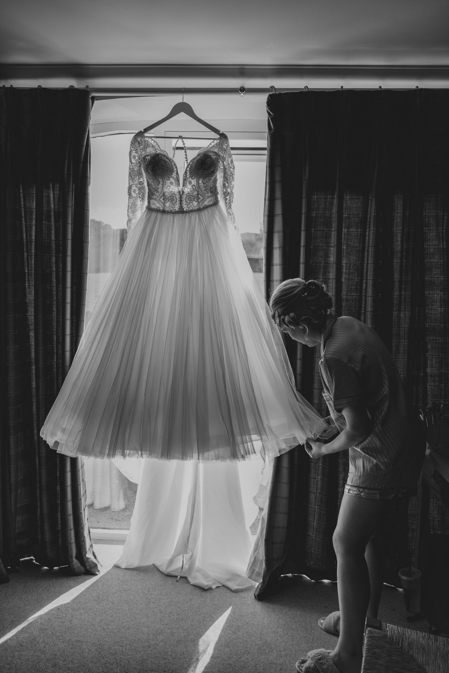 Black and white photo of a bride admiring her lace wedding gown hanging by a window.