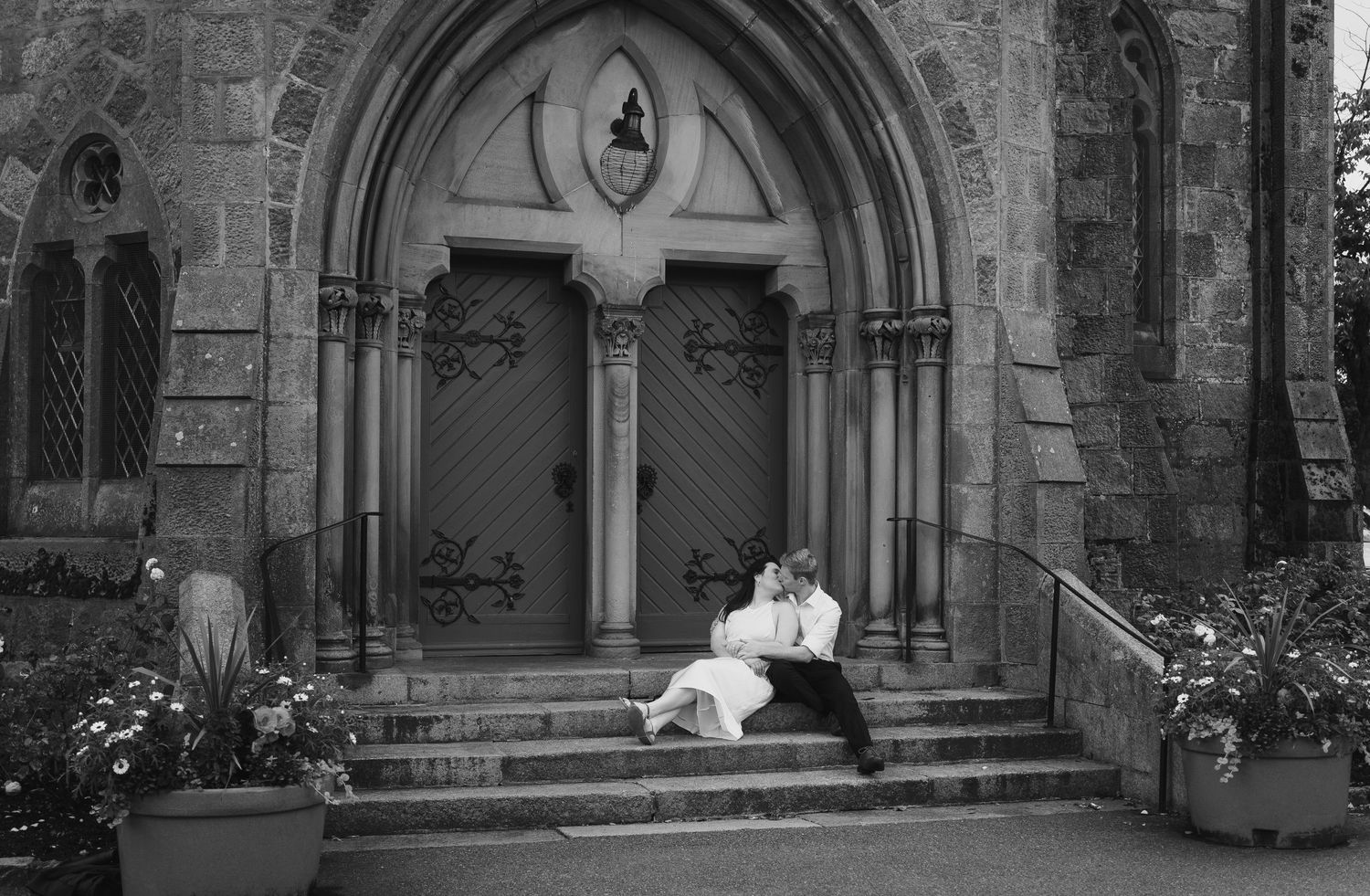 Couple sitting closely on stone steps of a grand Gothic church entrance with ornate wooden doors.