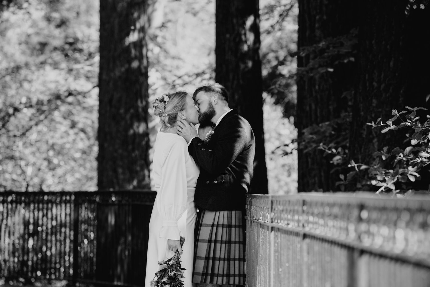 Black and white romantic photo of bride and groom kissing near tall trees in woodland.