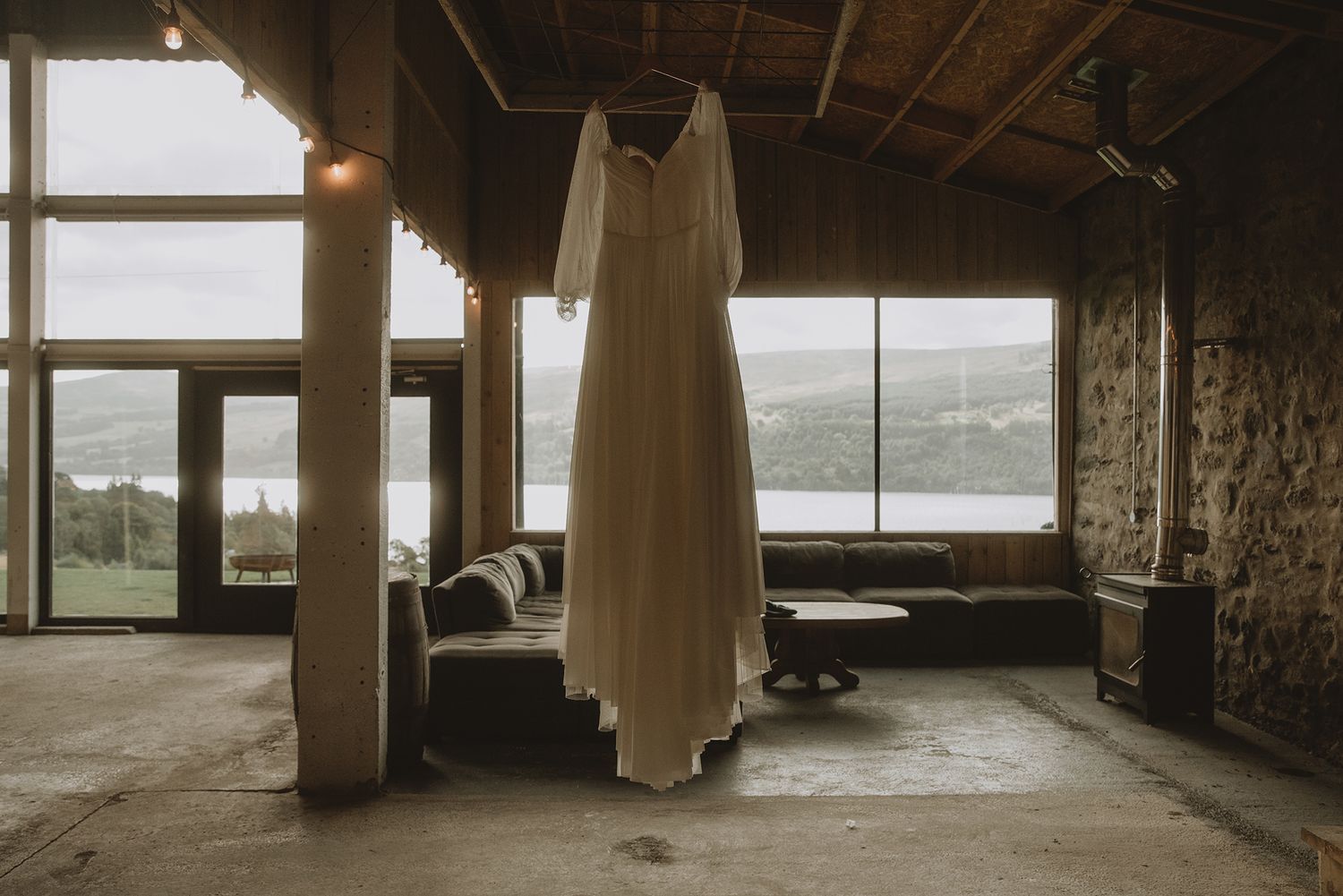 Elegant white wedding dress hanging in a rustic stone venue with panoramic loch views through windows.