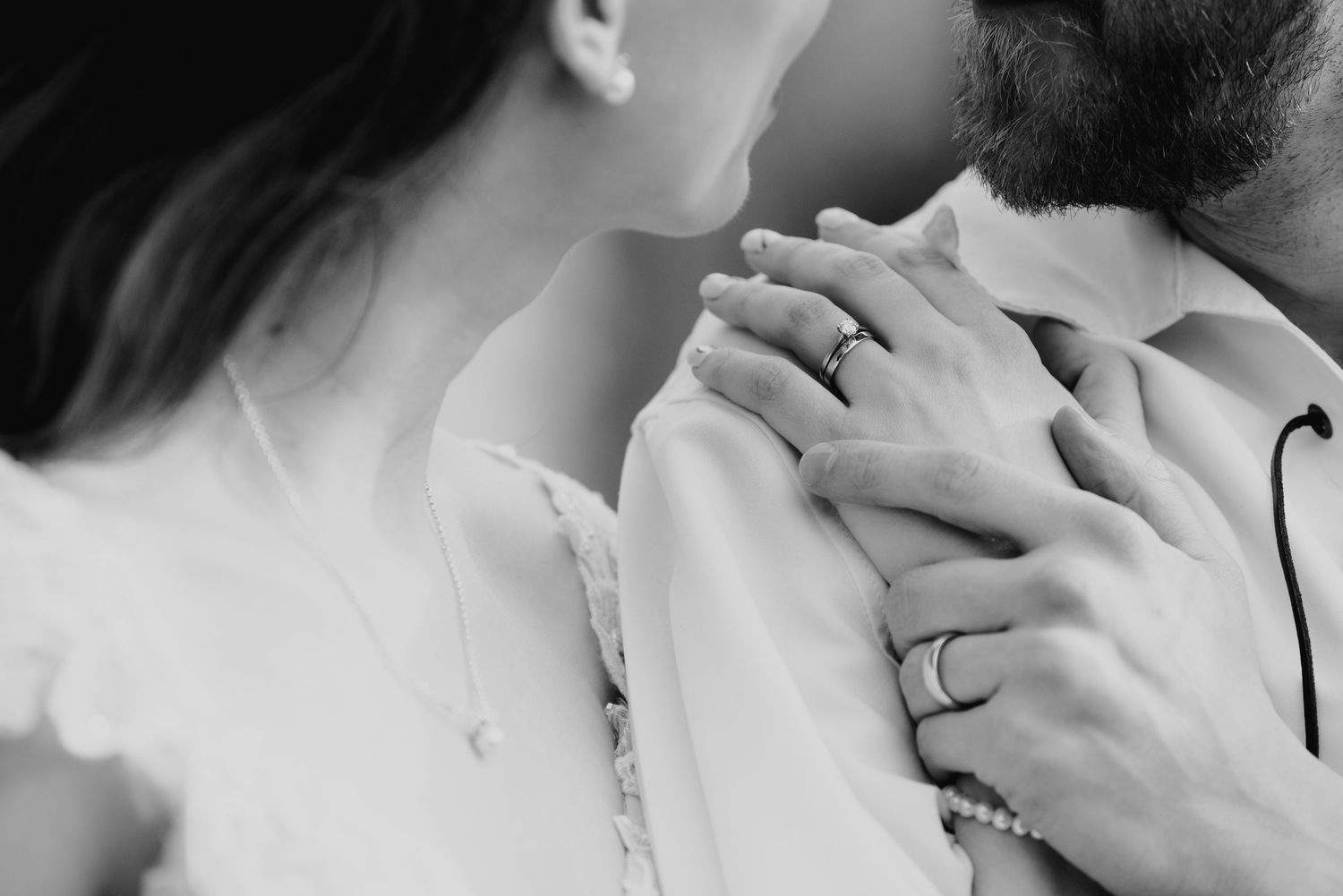 Black and white close-up of bride and groom embracing, showing wedding rings and delicate jewelry details.
