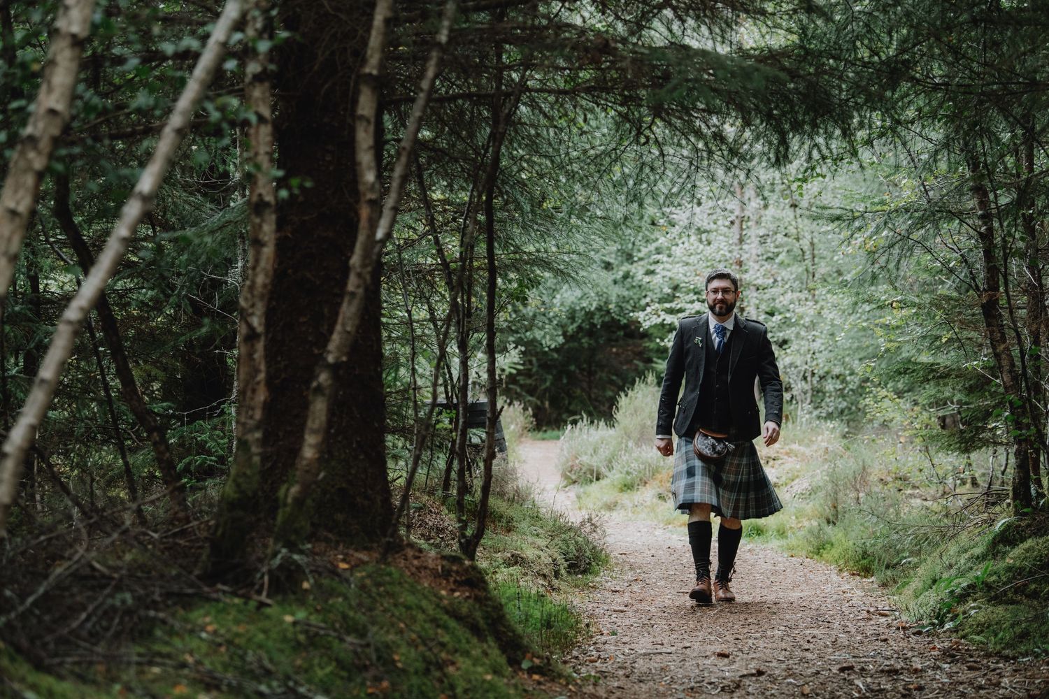 Groom in traditional Scottish kilt walks through lush forest woodland path.