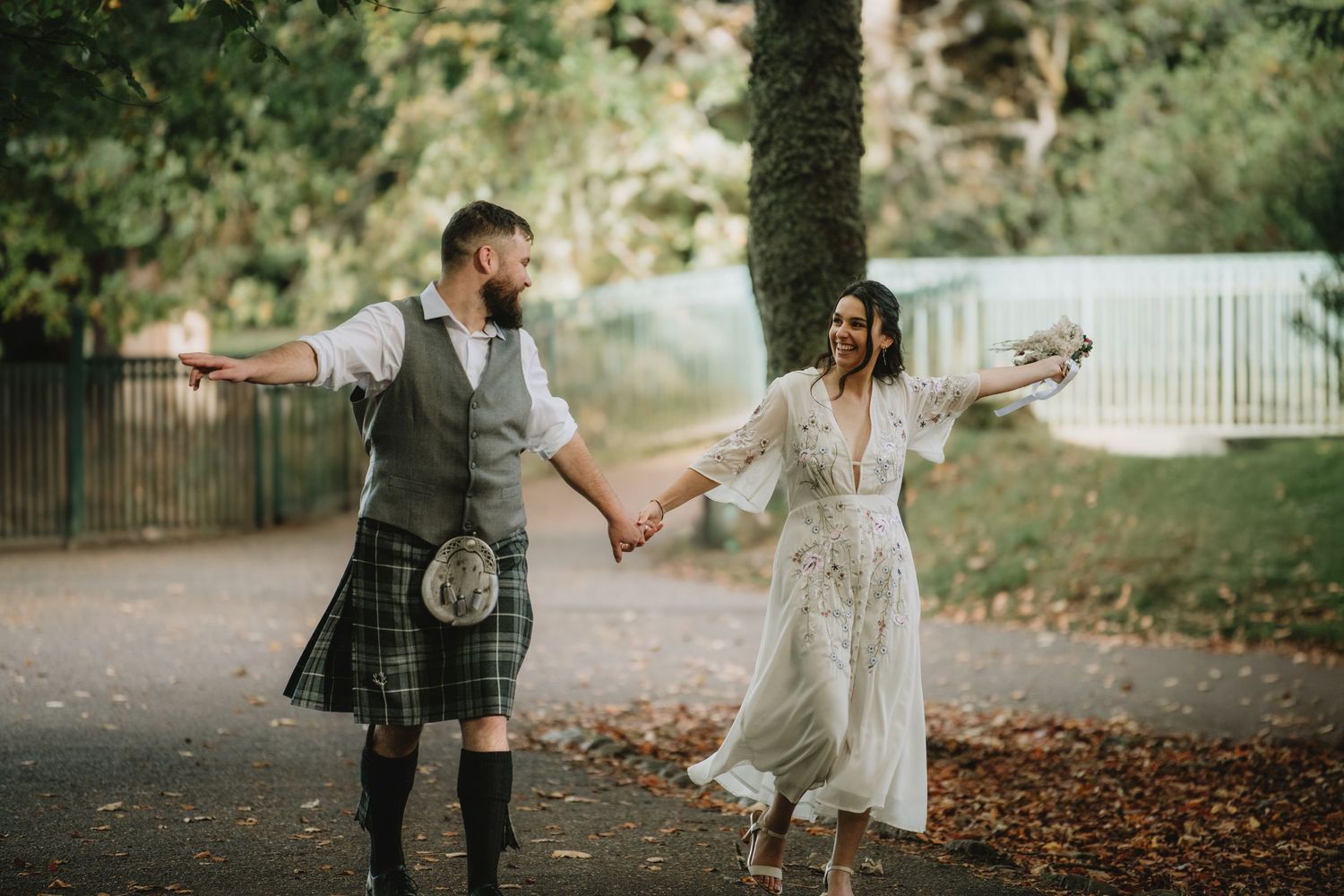 Joyful bride and groom in Scottish kilt dance hand-in-hand along tree-lined path in autumn park.