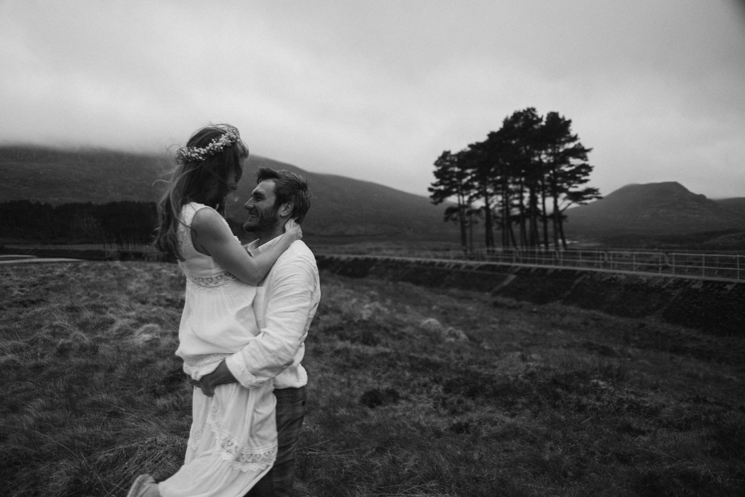 Black and white wedding photo of groom lifting bride in a scenic highland setting with trees.