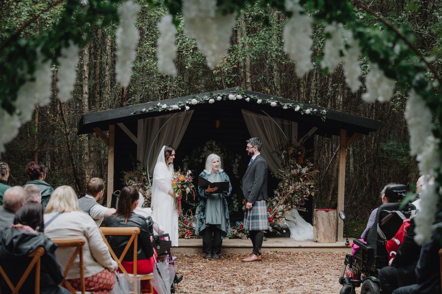 Couple exchanges vows under decorated woodland gazebo with floral arch during outdoor forest wedding ceremony.