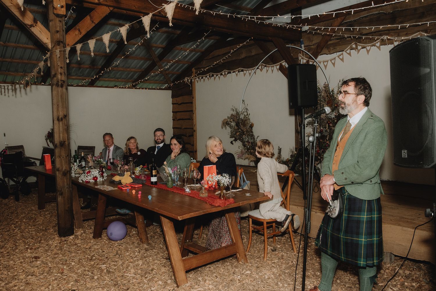 Wedding guest in kilt giving speech to seated guests in rustic barn venue with bunting.