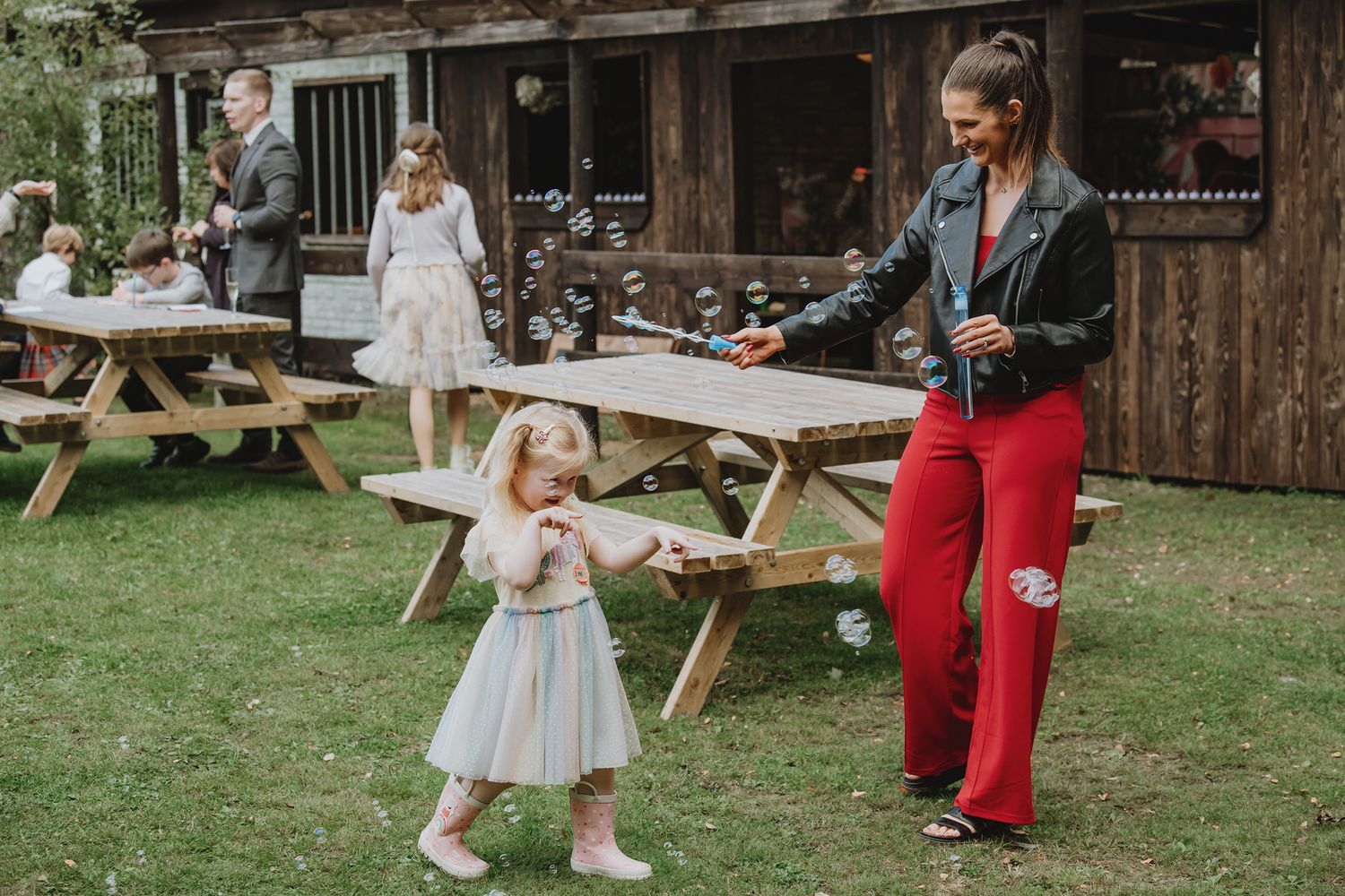A woman in red trousers blows bubbles for a young girl in a white dress at an outdoor wedding.