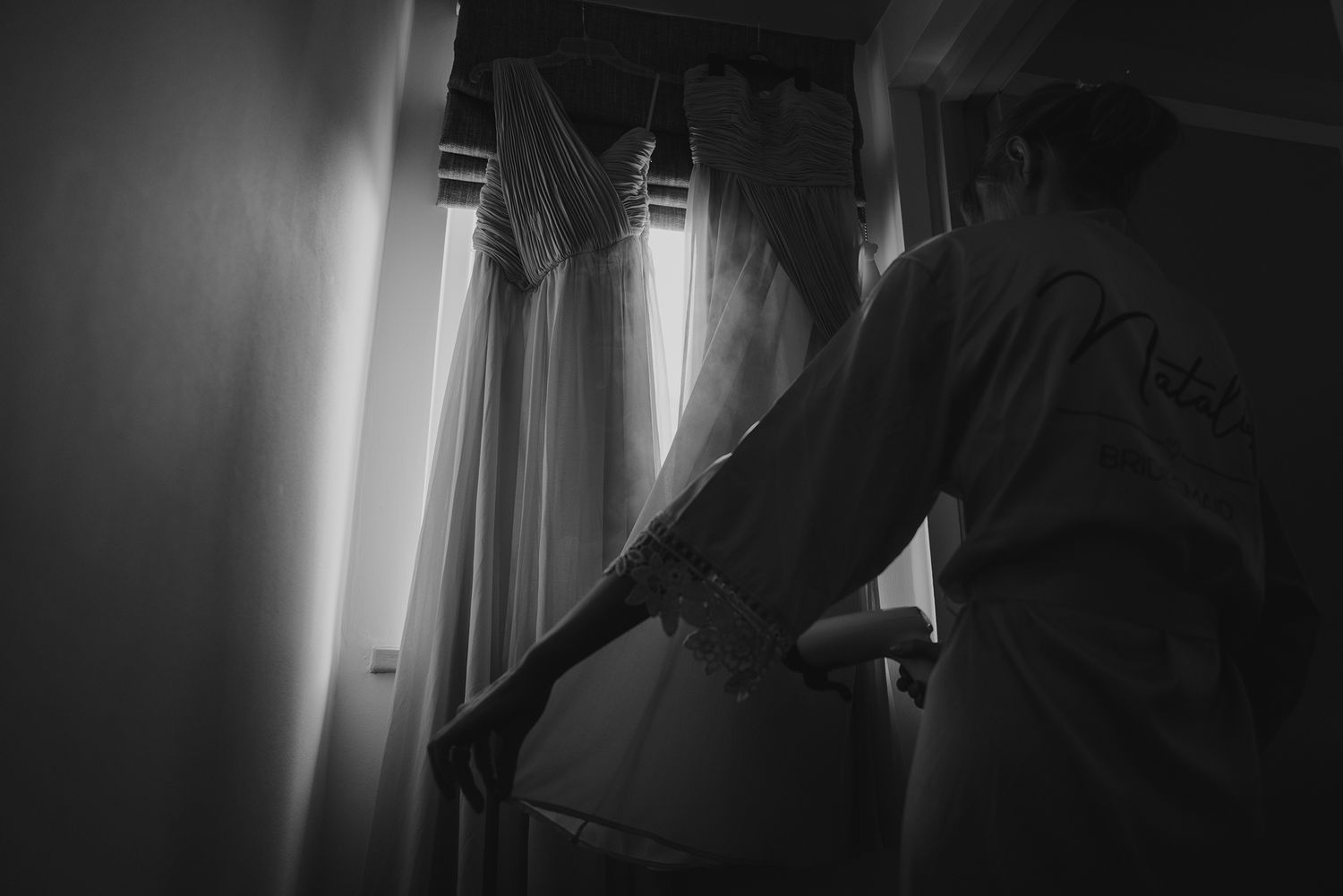 Black and white photo of a bride steaming her wedding dress near a window before the ceremony.