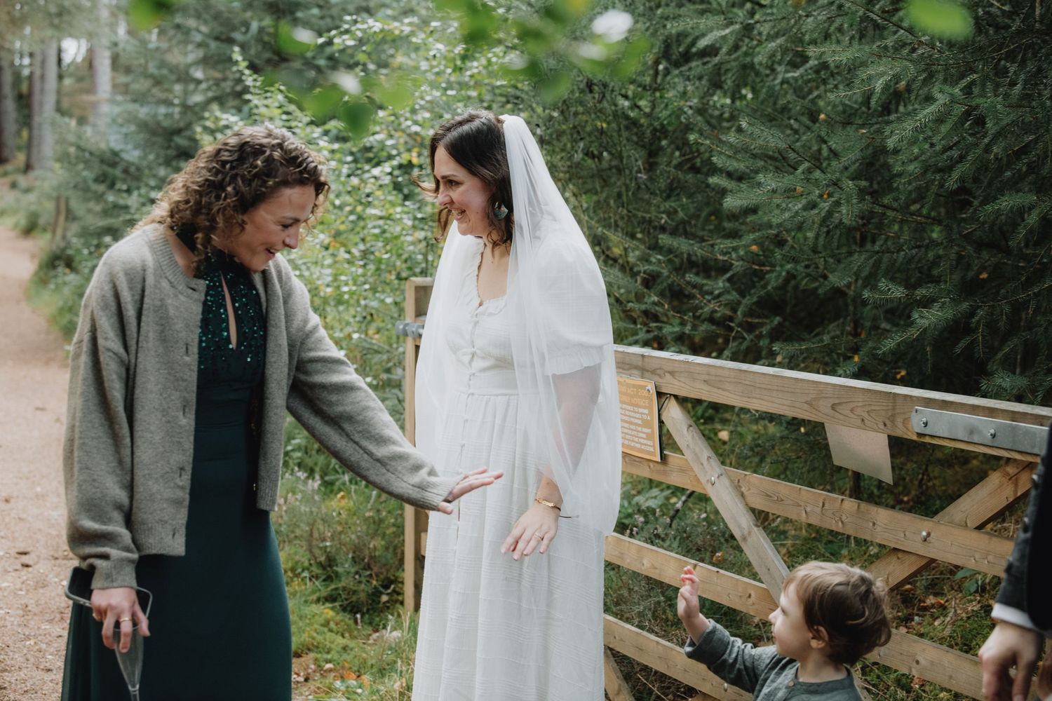 Bride in white wedding veil chats with female guest by wooden gate as child reaches up in forest.