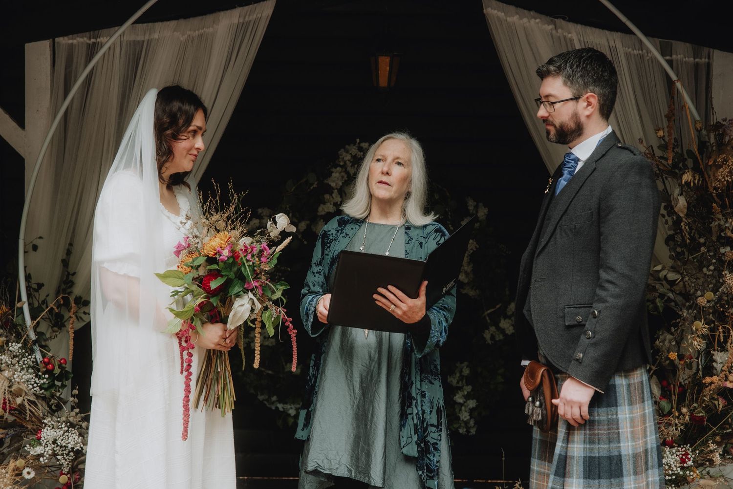 Bride with veil holds vibrant bouquet beside officiant and bearded groom in kilt at forest ceremony.