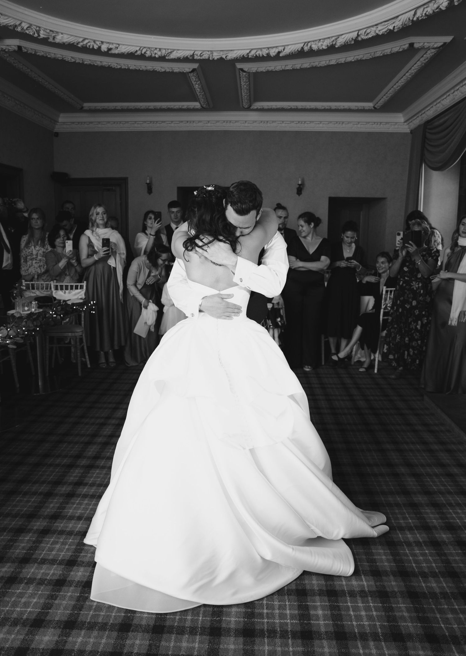 Black and white photo of bride and groom sharing an intimate first dance on a tartan floor before guests.