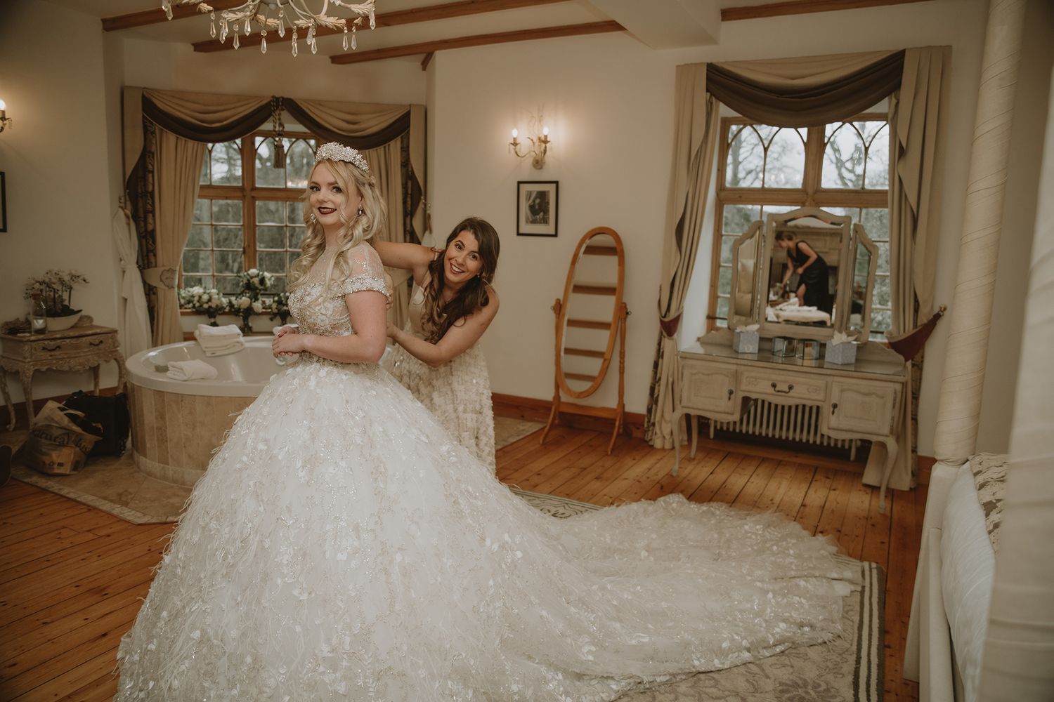 Bride in elegant lace ball gown being helped by bridesmaid in a vintage-styled room with ornate decor.