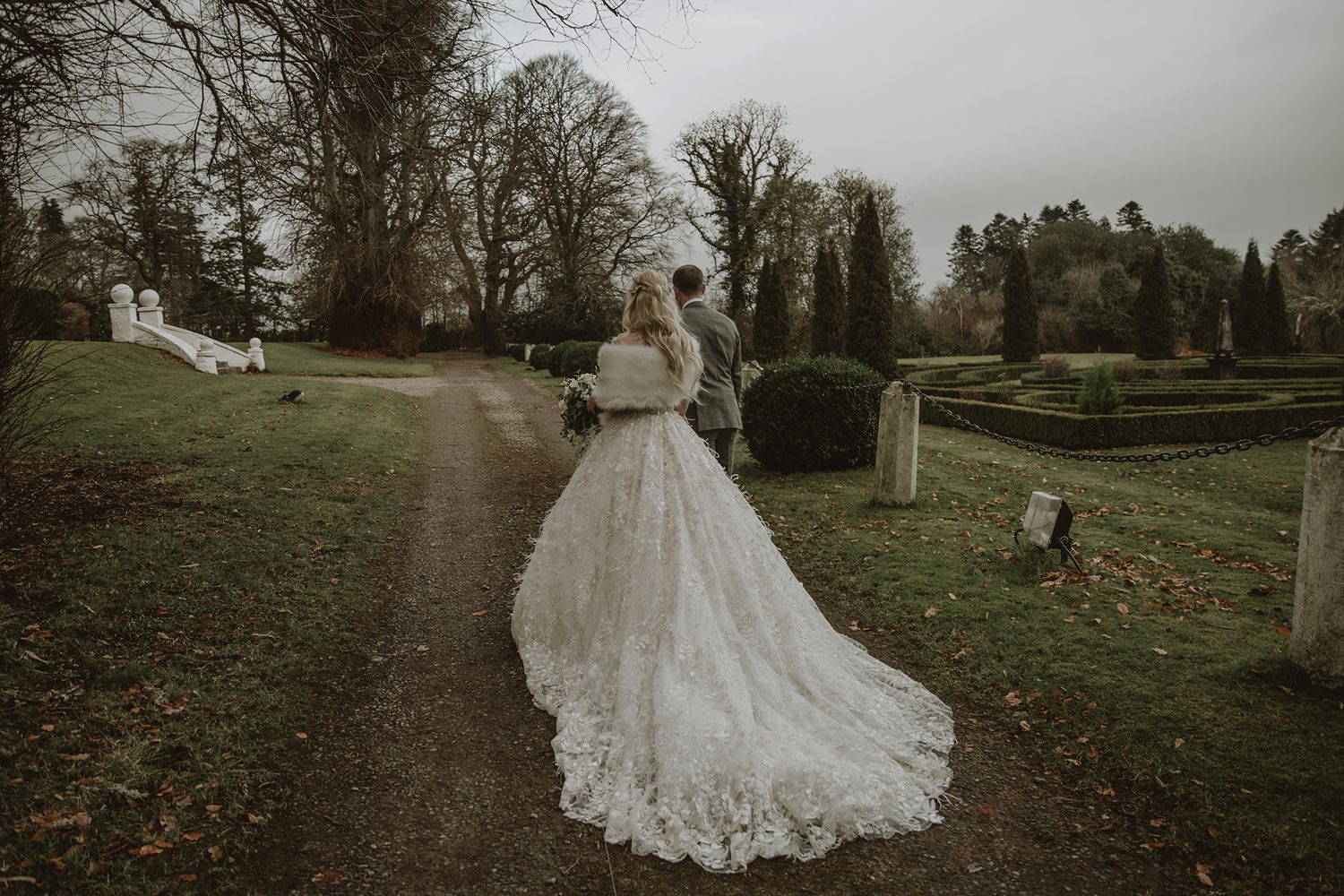 Bride in lace gown with long train walks with groom through scenic garden path on overcast winter wedding day.