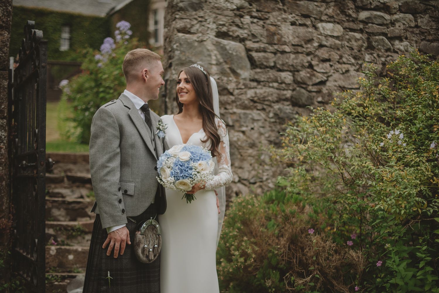 Bride and groom holding blue hydrangea bouquet beside stone wall in Scottish wedding attire including kilt.