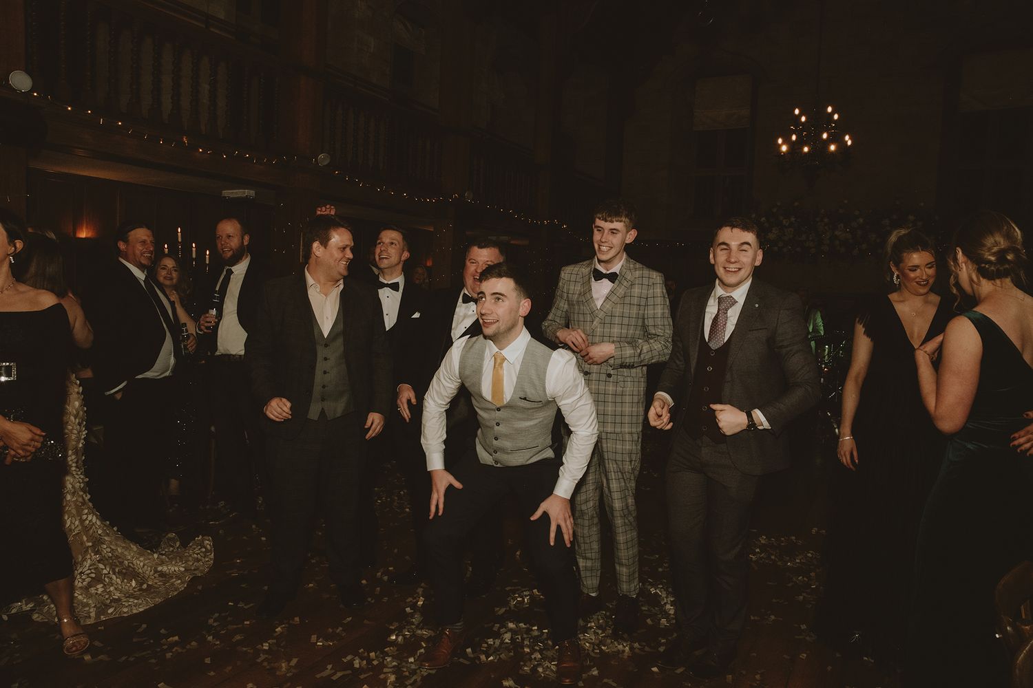 Groomsmen dancing energetically at a wedding reception in a dimly lit elegant venue.