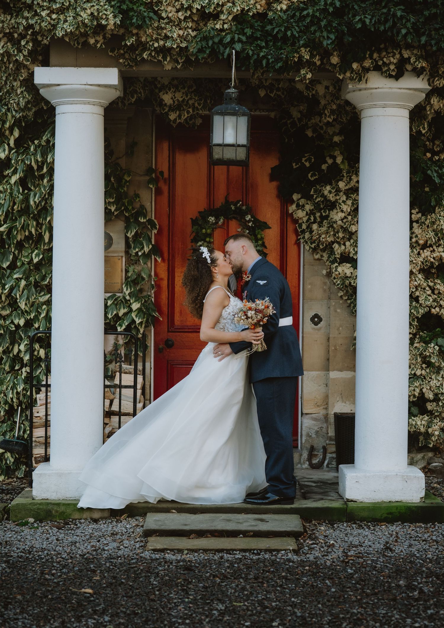 Bride and groom share romantic kiss at ivy-covered venue entrance with red door and white columns.