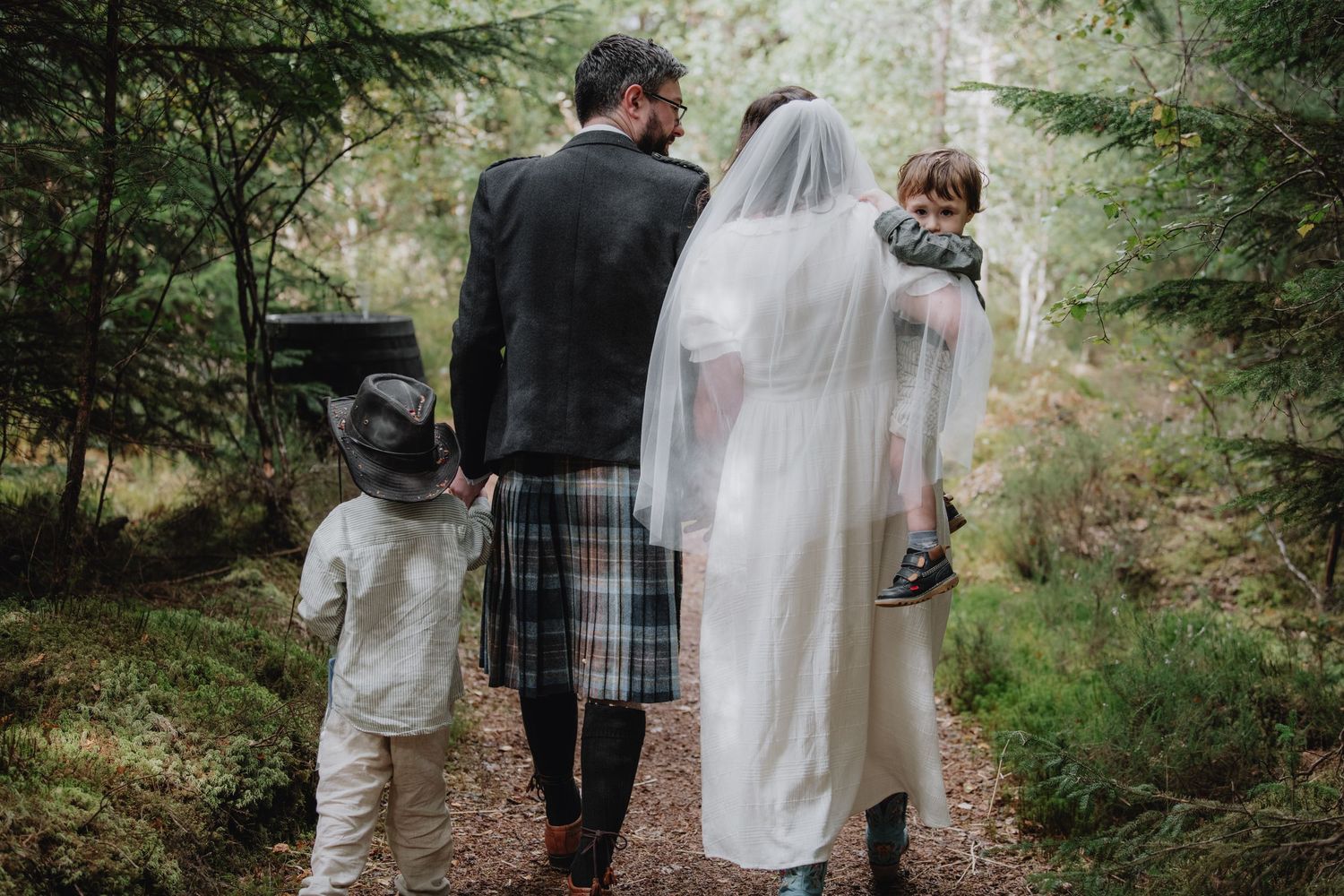 A wedding couple with two children walk hand-in-hand through a lush forest path, groom wearing a tartan kilt.