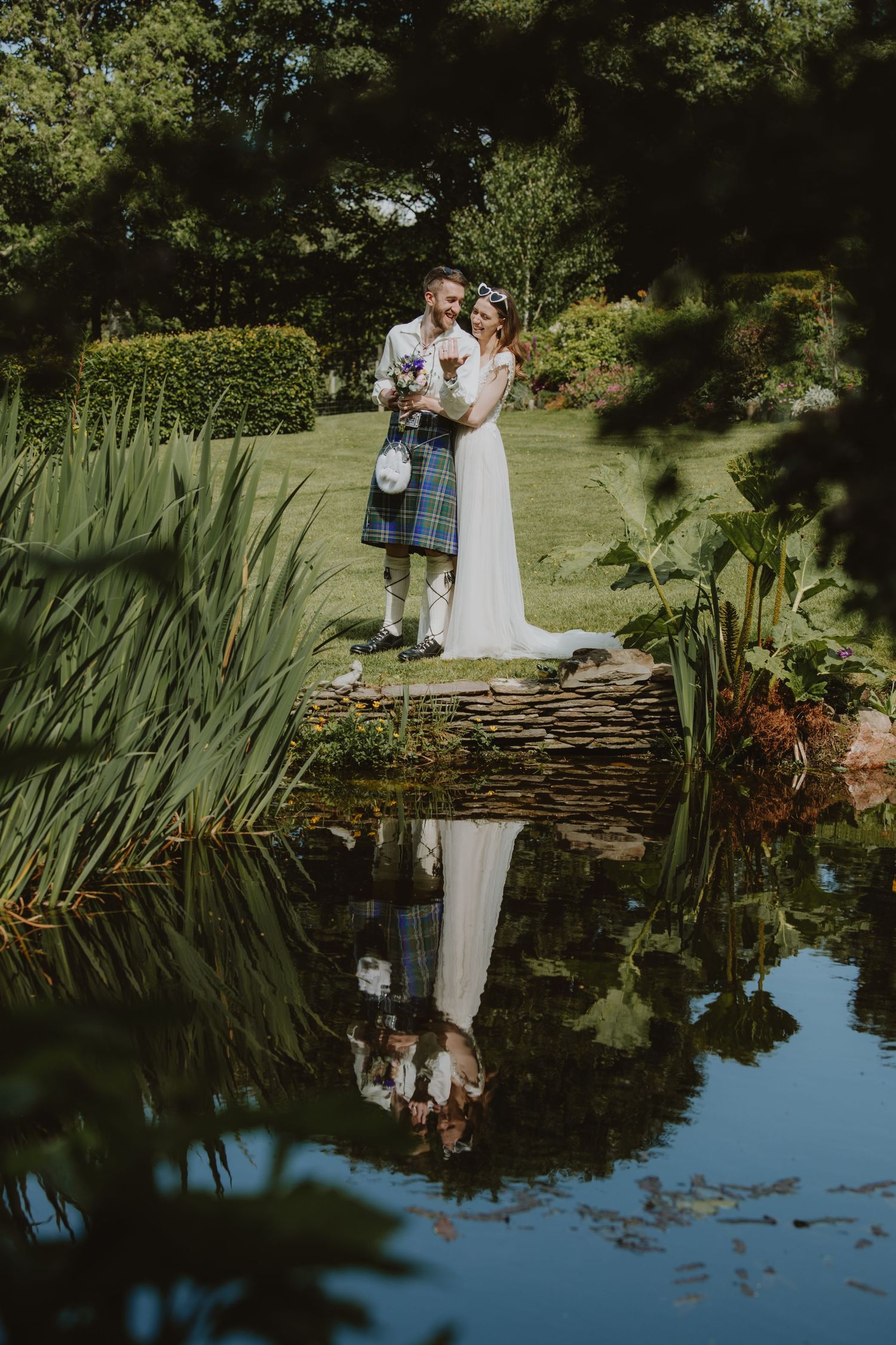 Bride and groom reflect in a garden pond as they stand on a stone bridge surrounded by lush greenery.