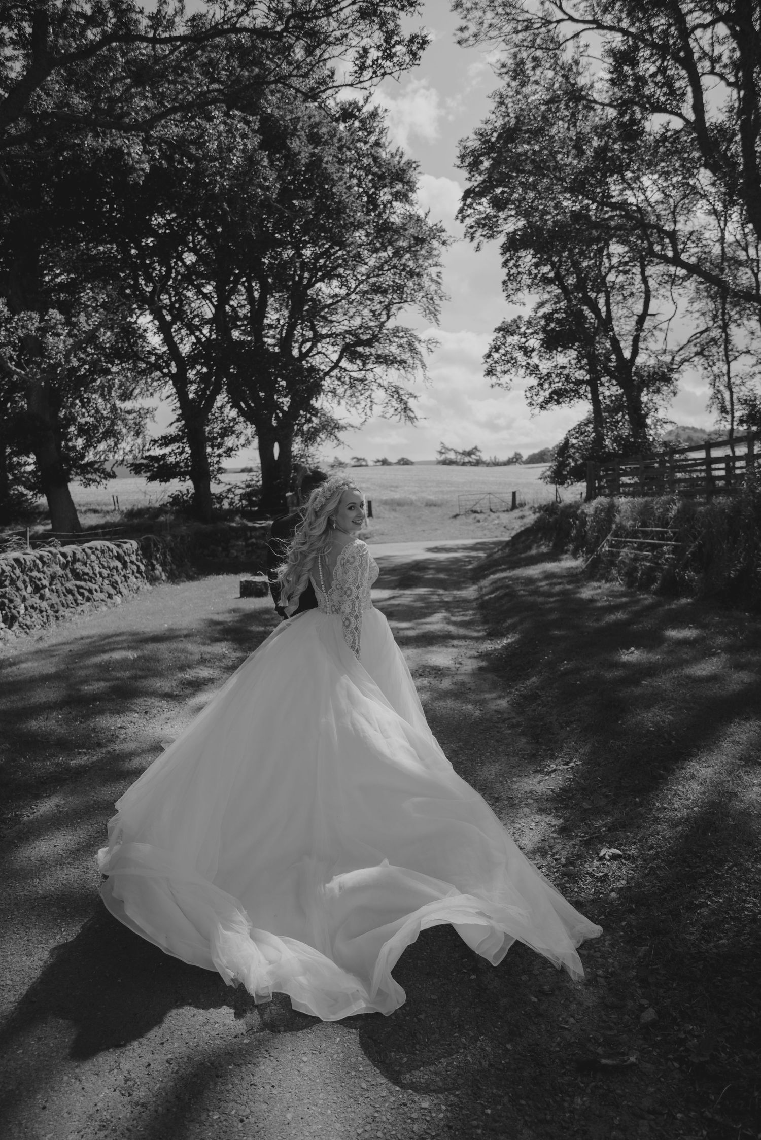 Bride in flowing white gown walking on a tree-lined path in black and white.