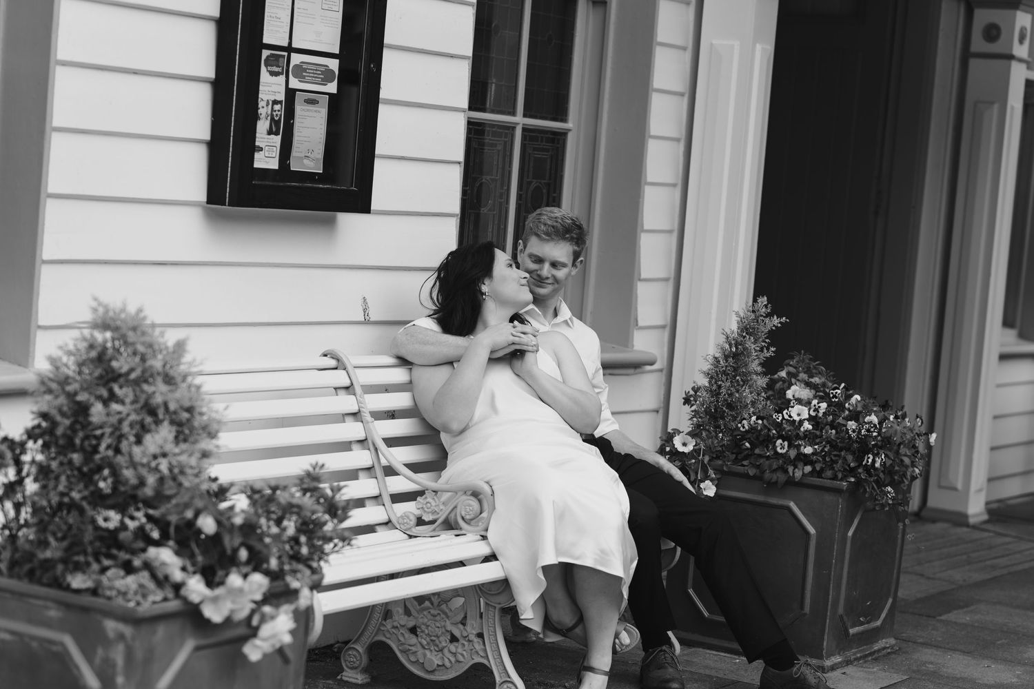 Happy couple sharing a kiss on a white wooden bench outside a charming building with flowers.