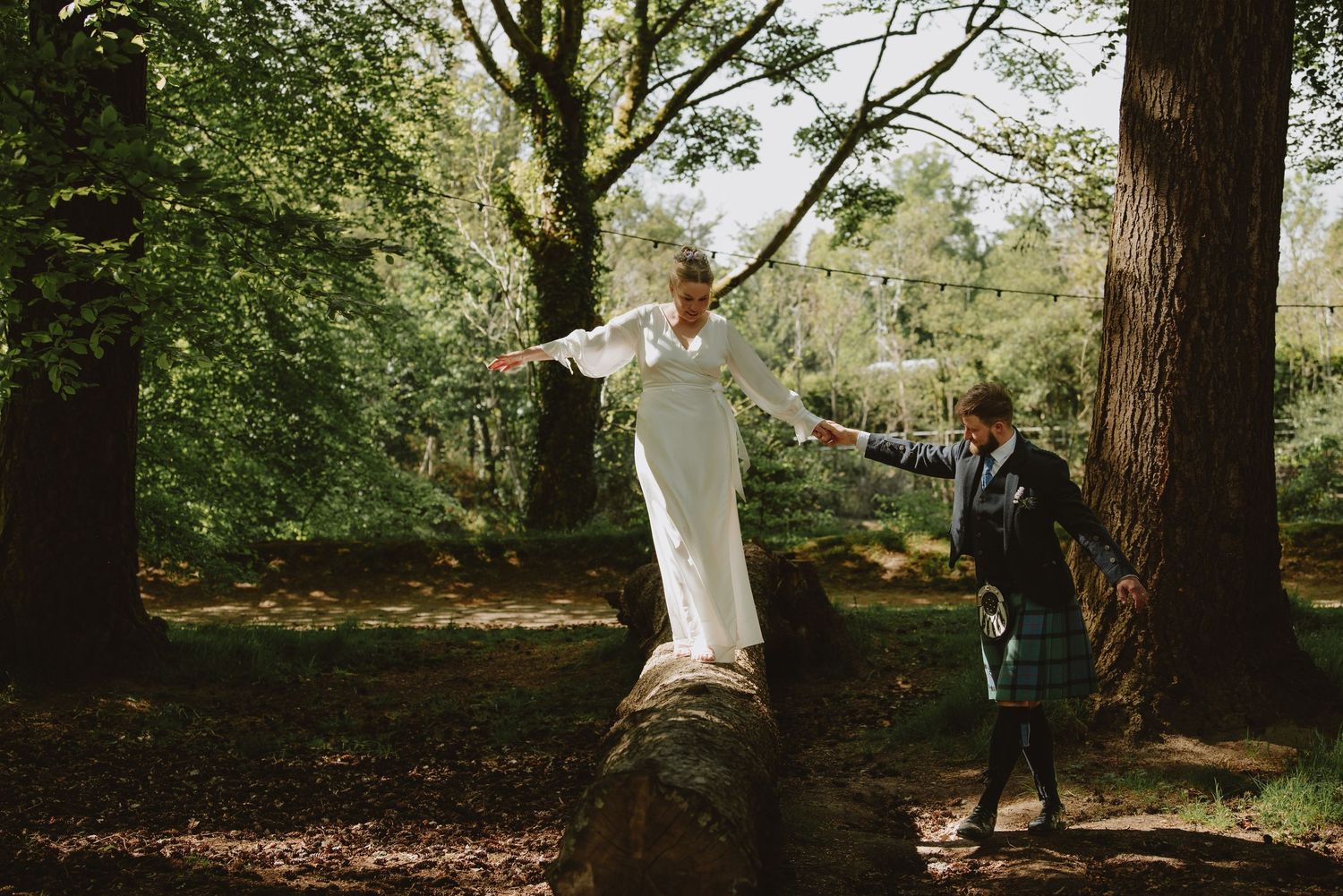 Colorful outdoor photo of bride balancing on fallen log as groom steadies her in forest.