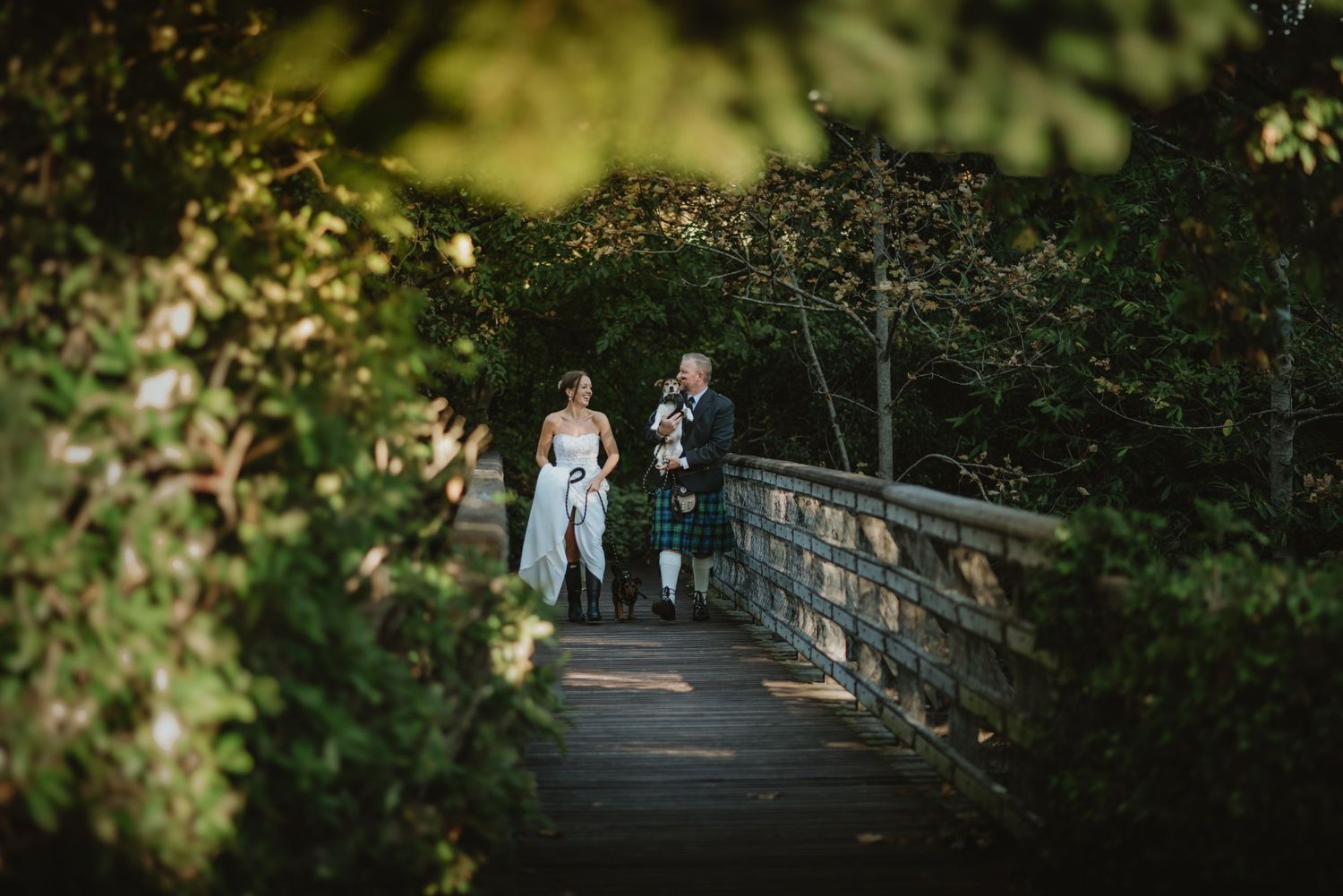 Bride and groom in Scottish kilt walk hand in hand on a wooden bridge surrounded by lush green foliage.