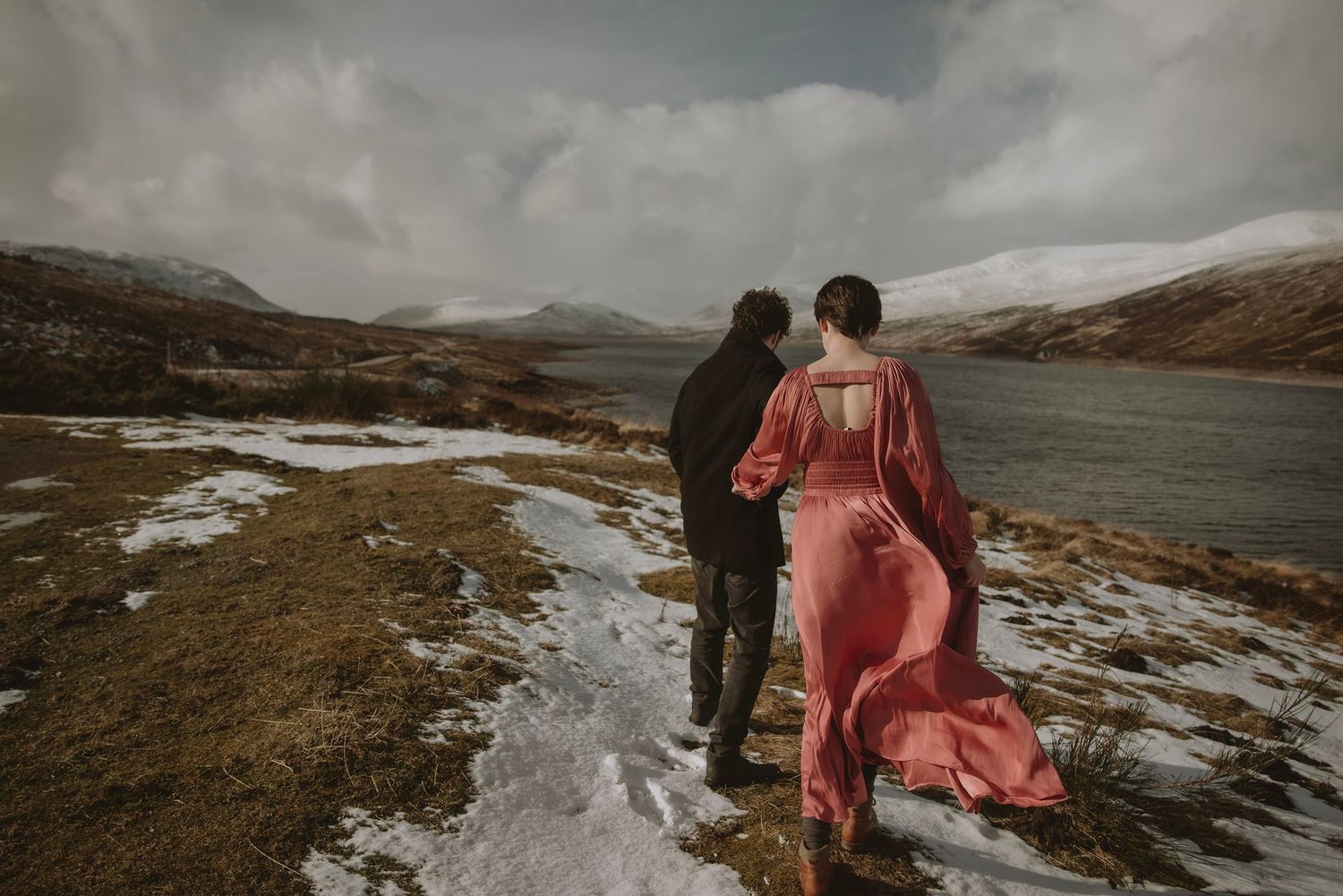Couple walking along a snowy highland loch shore, woman wearing a flowing red gown.