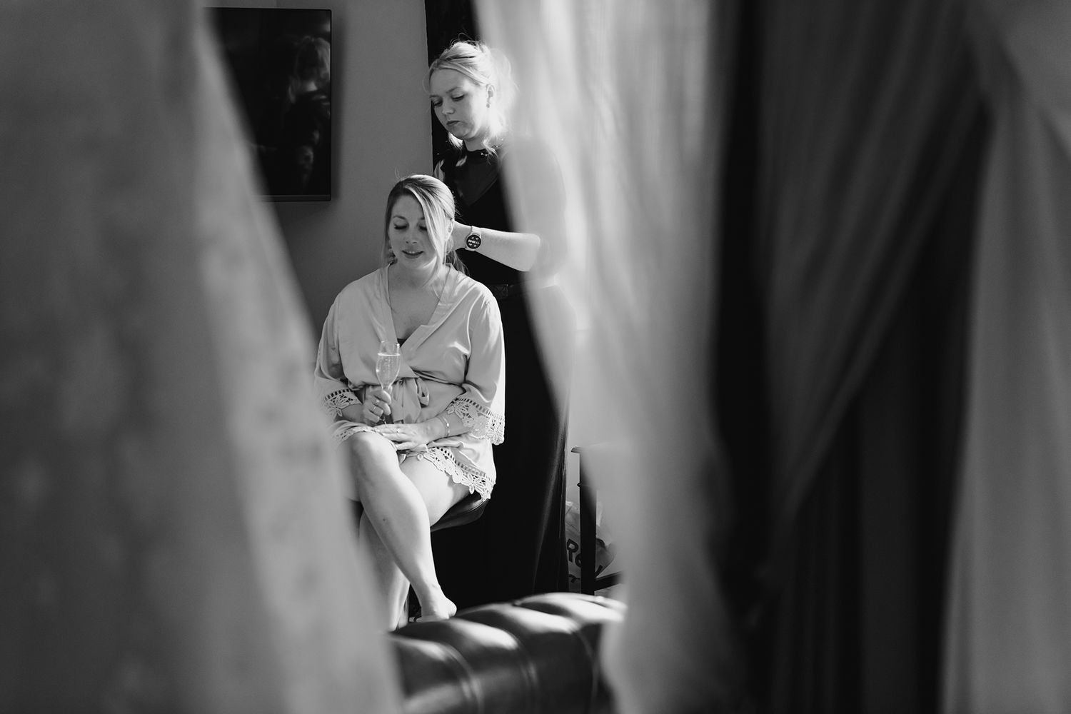 Black and white photo of bride in robe having hair styled before wedding ceremony.