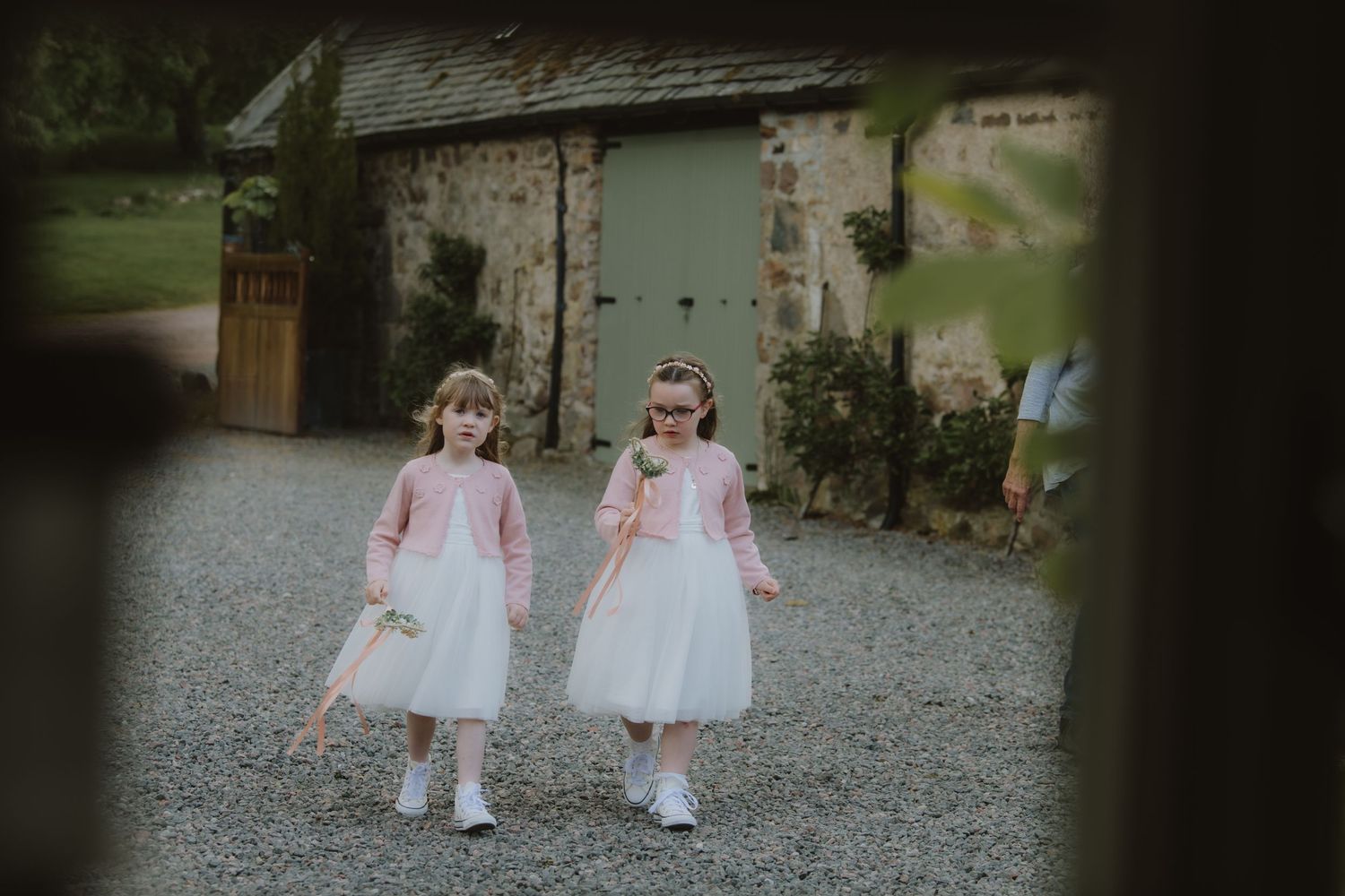 Two flower girls in white dresses and pink cardigans walk together on a gravel path at a rustic venue.