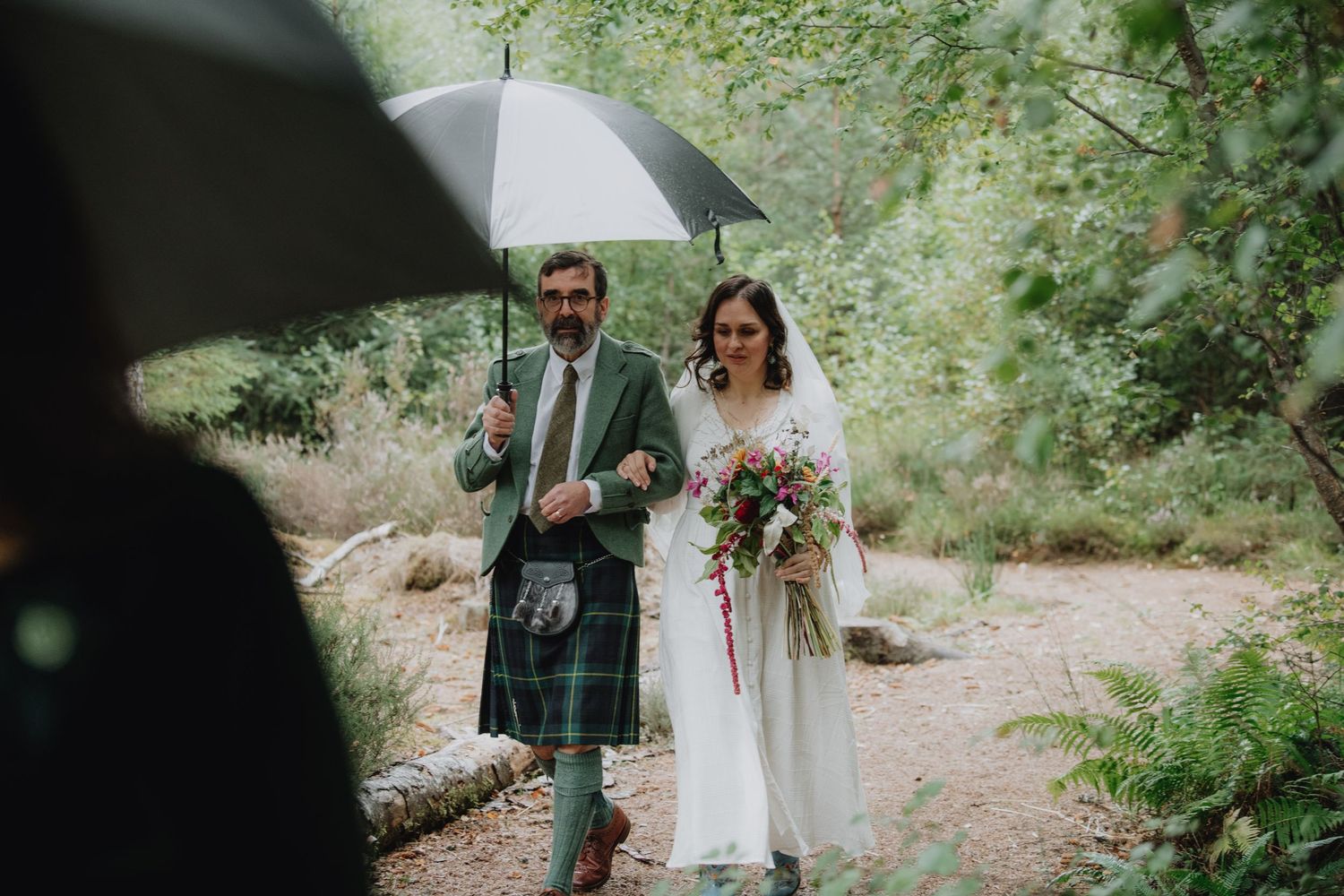 Groom in green tartan kilt walking bride in white gown along a rainy forest path with umbrella.