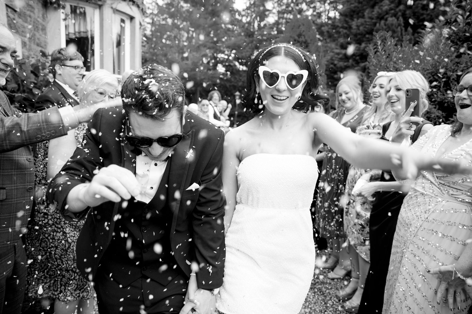 Black and white photo of smiling bride in heart sunglasses and groom walking through confetti with wedding guests.