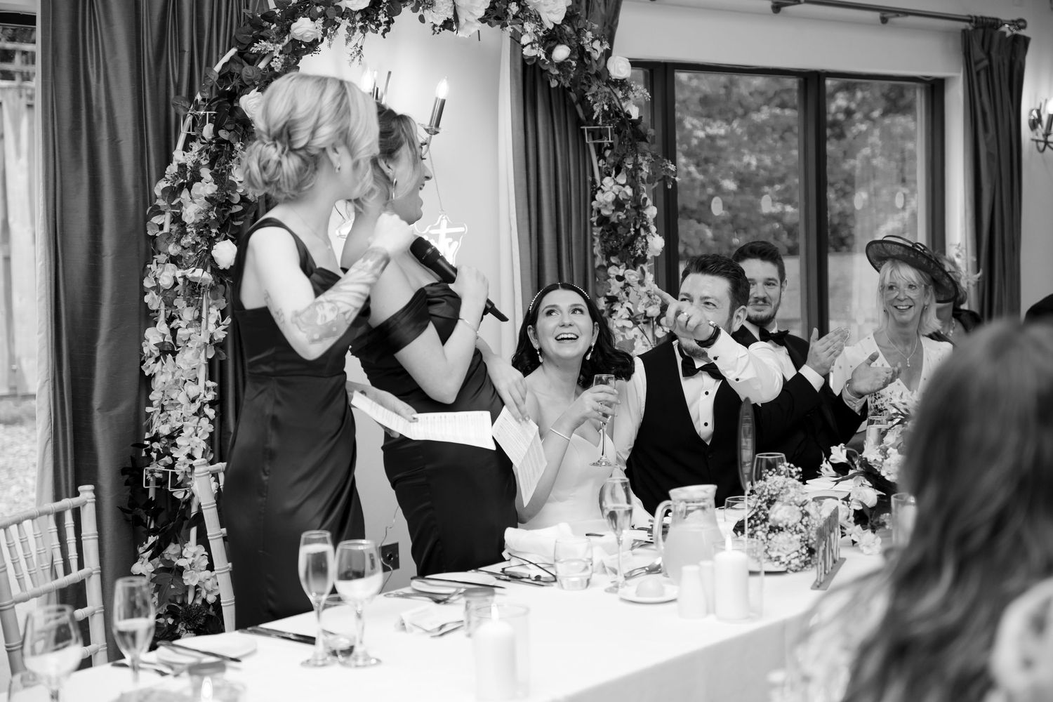 Black and white photo of wedding party laughing and celebrating during a lively speech at the reception table.